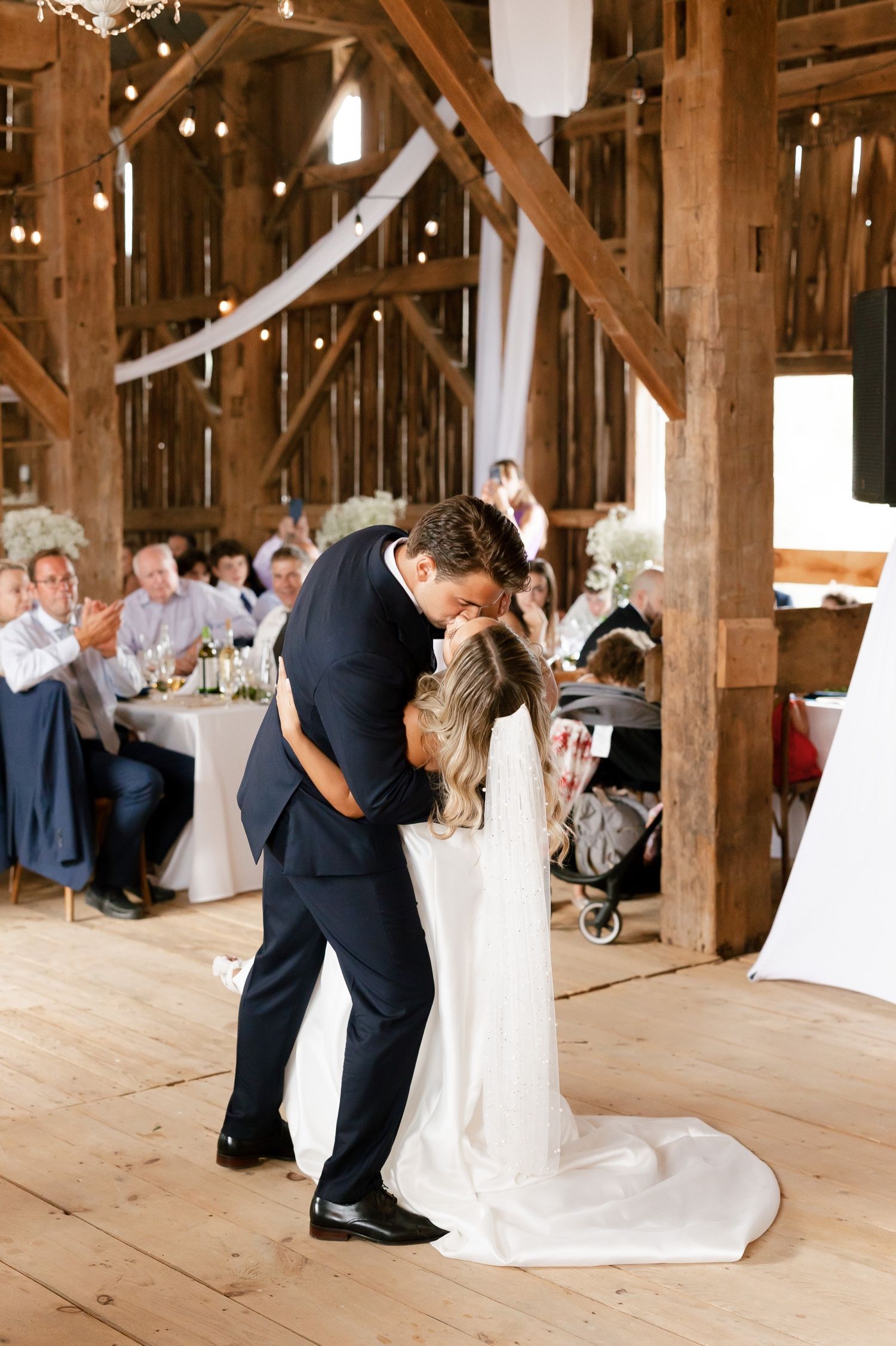 A tender embrace during a first dance in a rustic wooden barn with guests looking on.