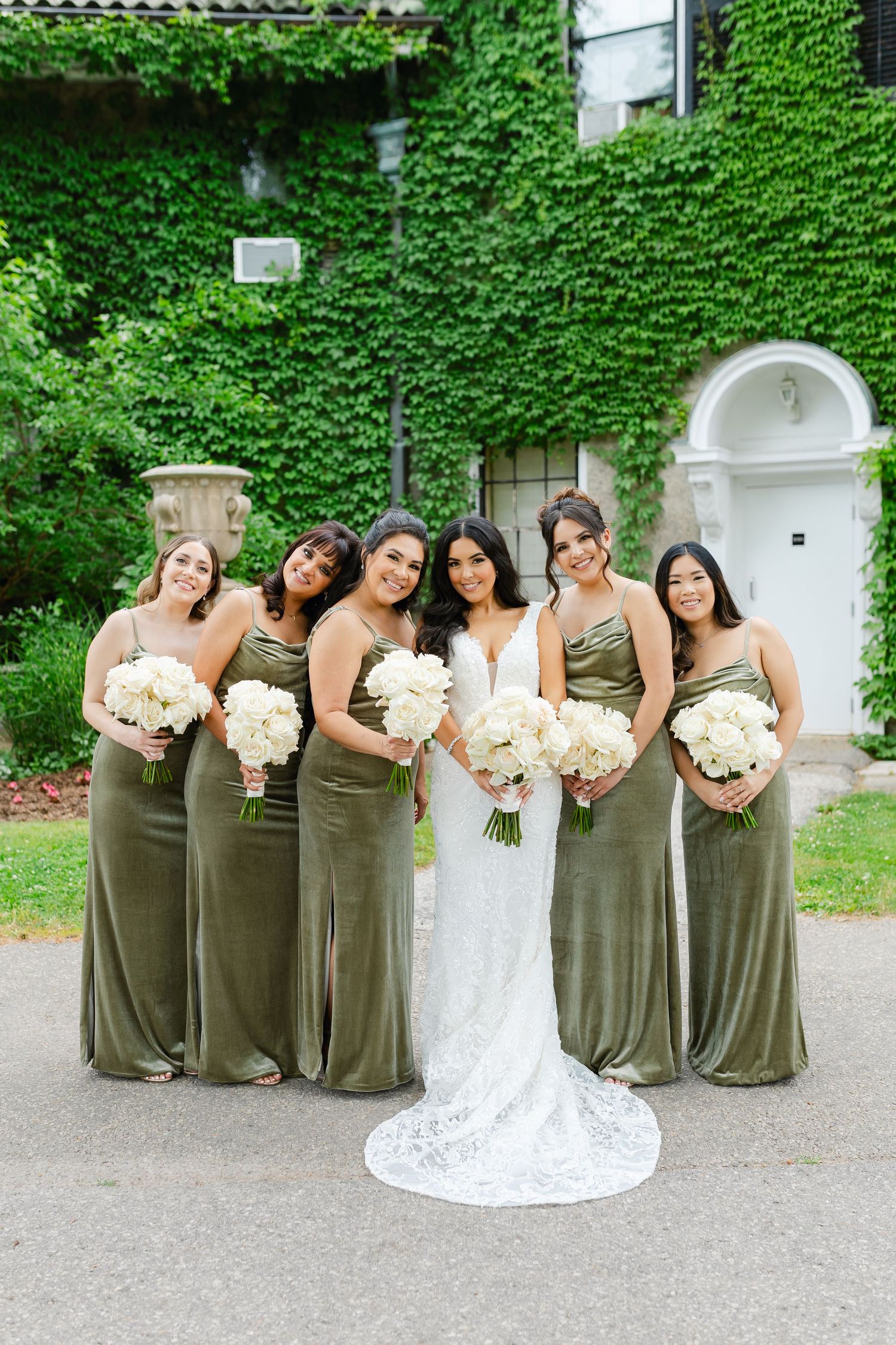 Bridesmaids in olive gowns pose with bride holding white bouquets against ivy-covered wall.