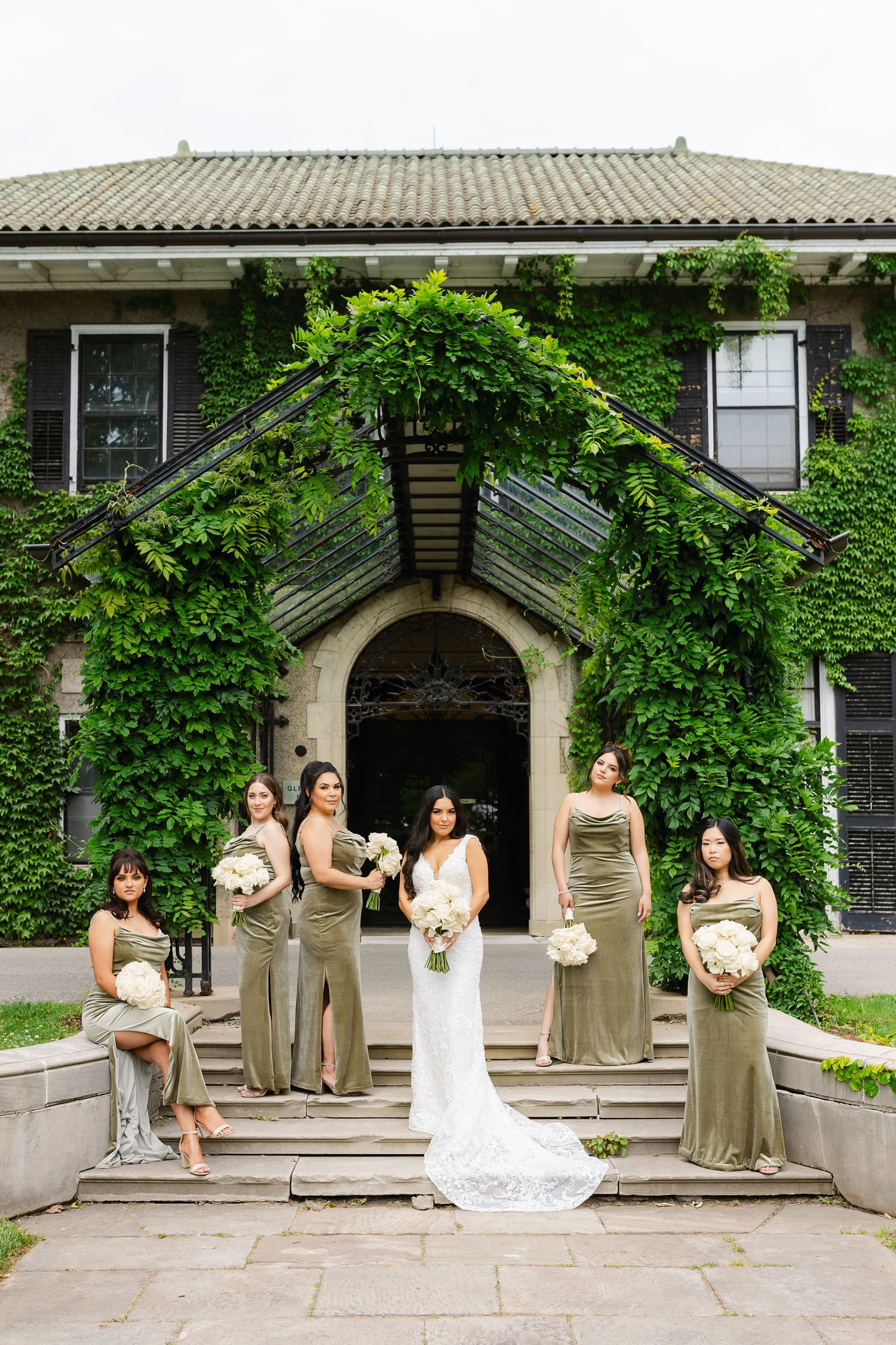 Bridal party in neutral gowns poses on stone steps of ivy-covered historic building.