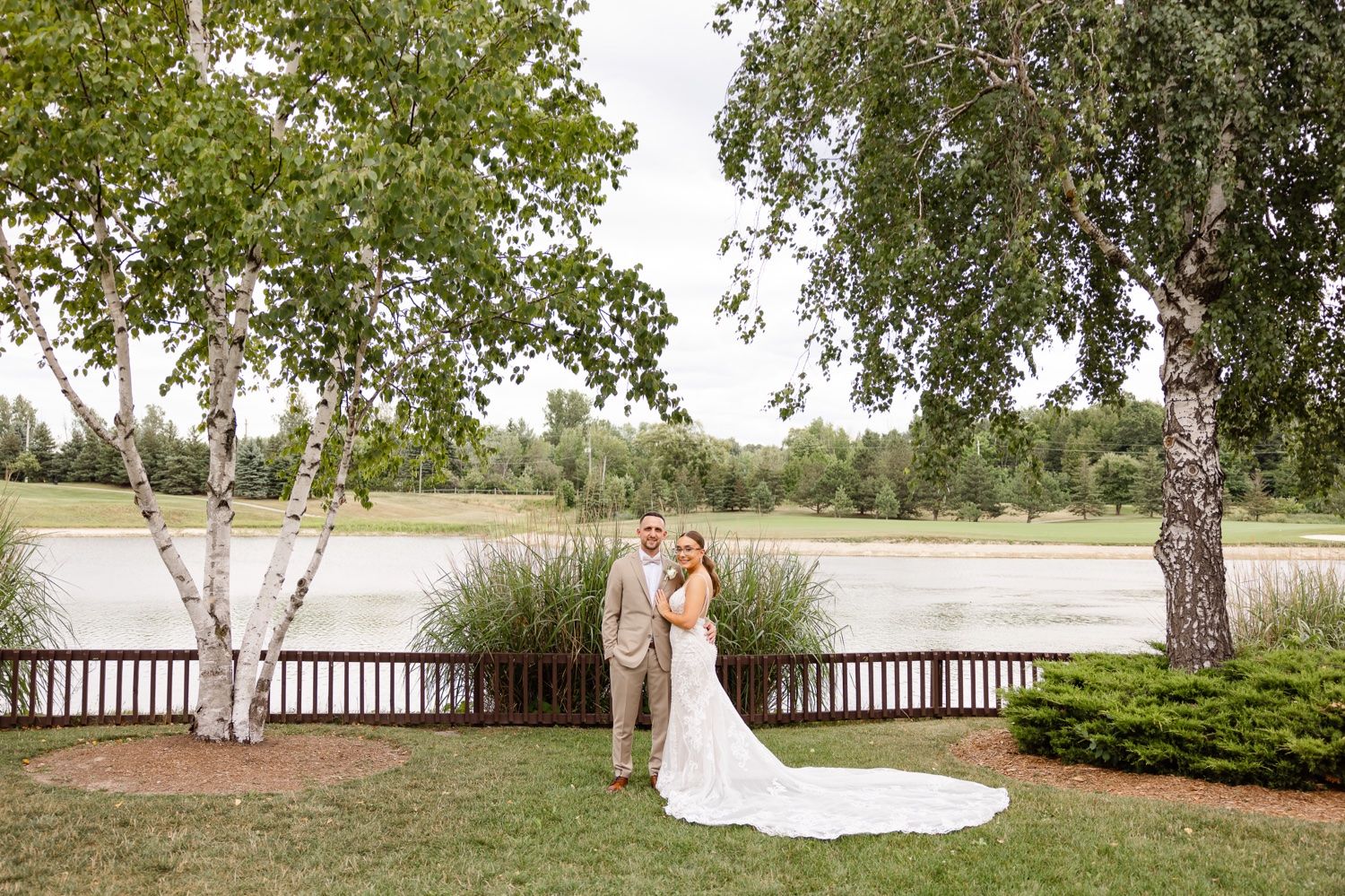 Scenic waterfront wedding portrait with couple standing between birch trees near a wooden deck.
