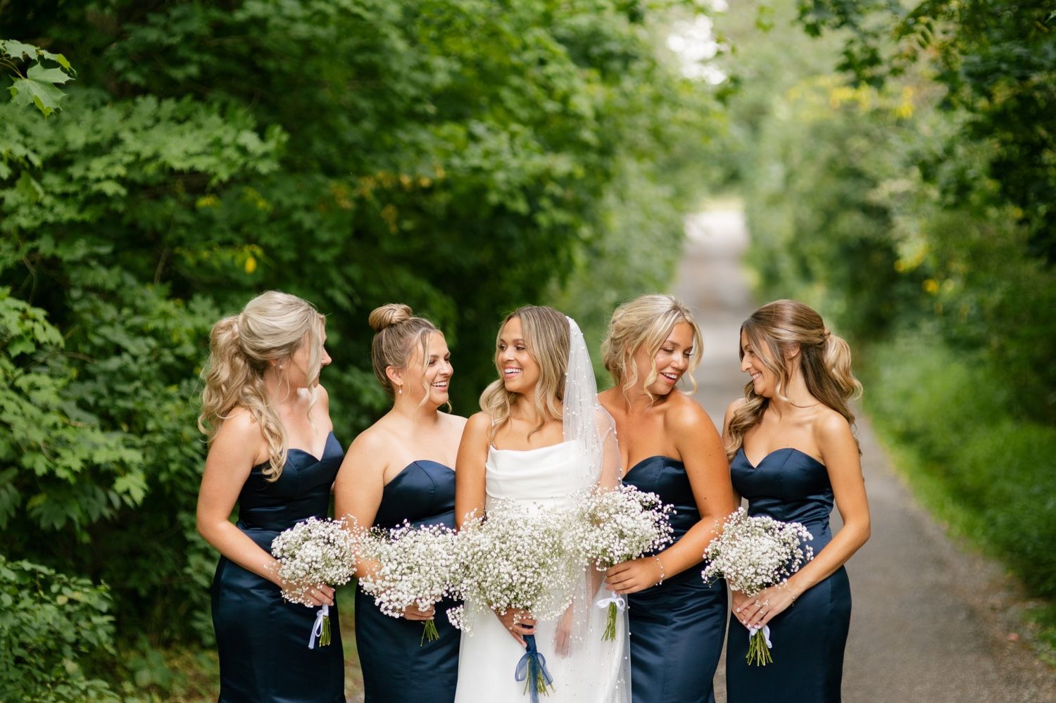 Bridal party poses joyfully together on rustic forest path with baby's breath bouquets.