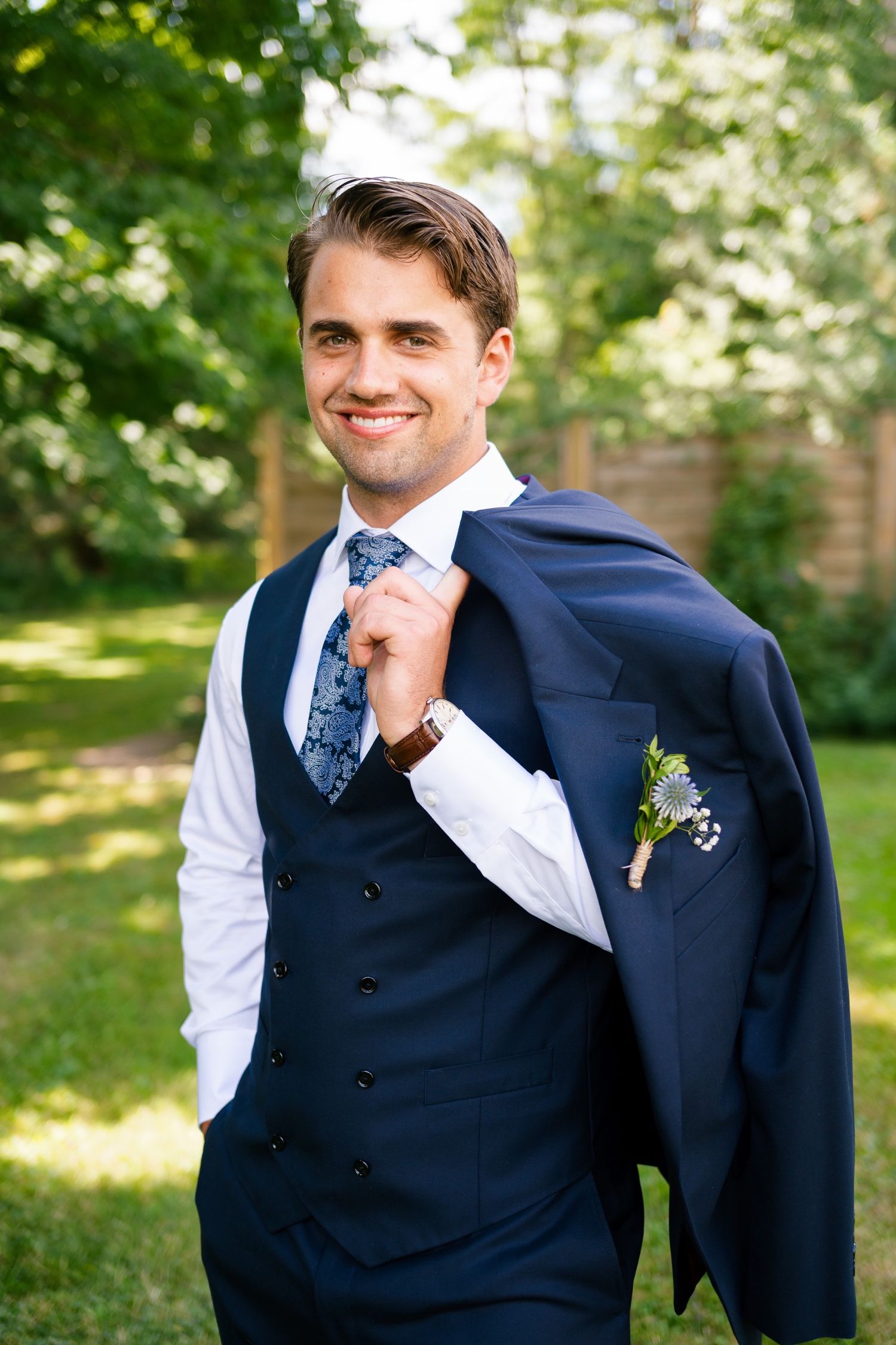 Stylish navy suit and vest with blue patterned tie and boutonniere in garden setting.