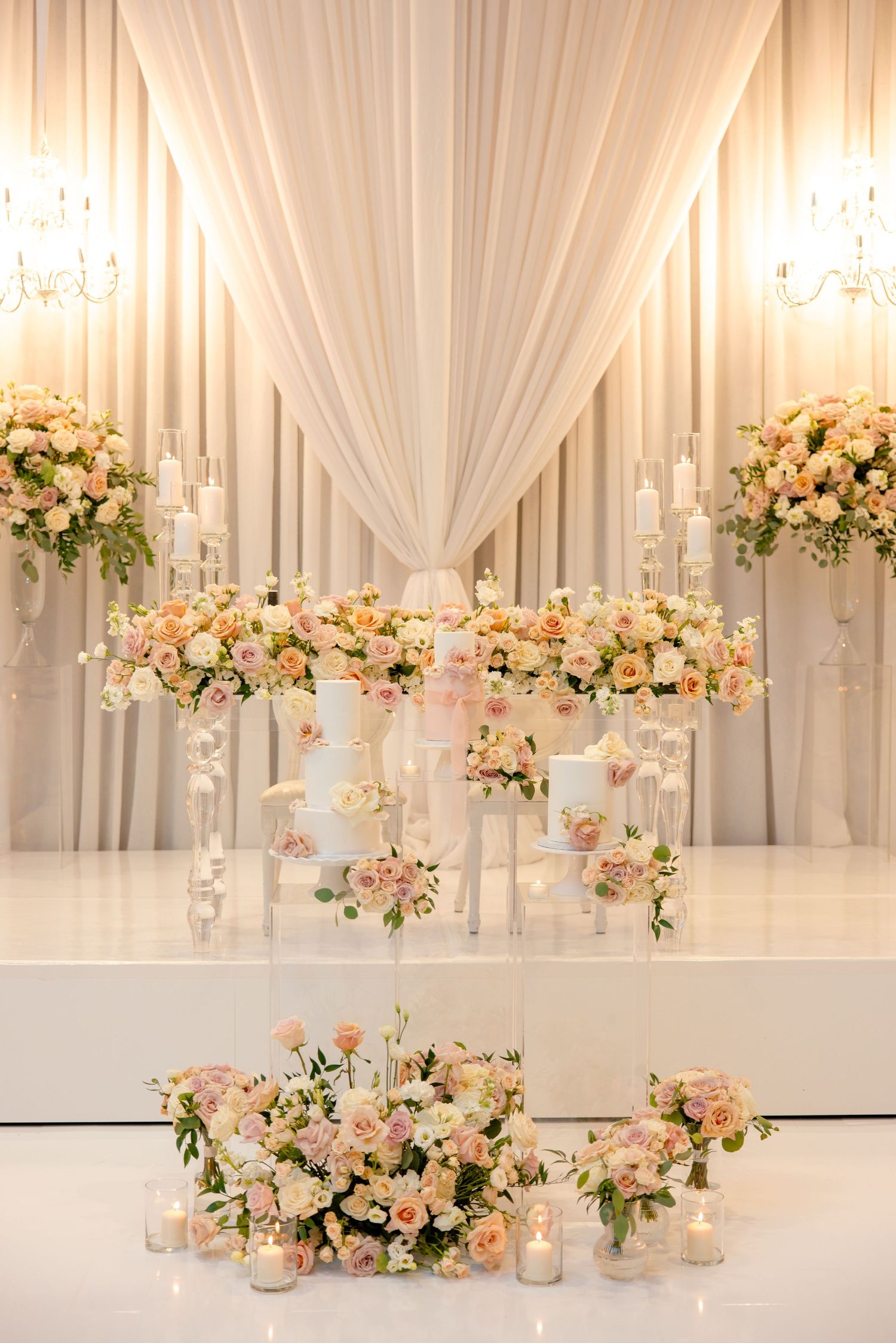 Elegant white draped backdrop with cascading floral arrangements and glowing candles creating a romantic wedding ceremony setting.