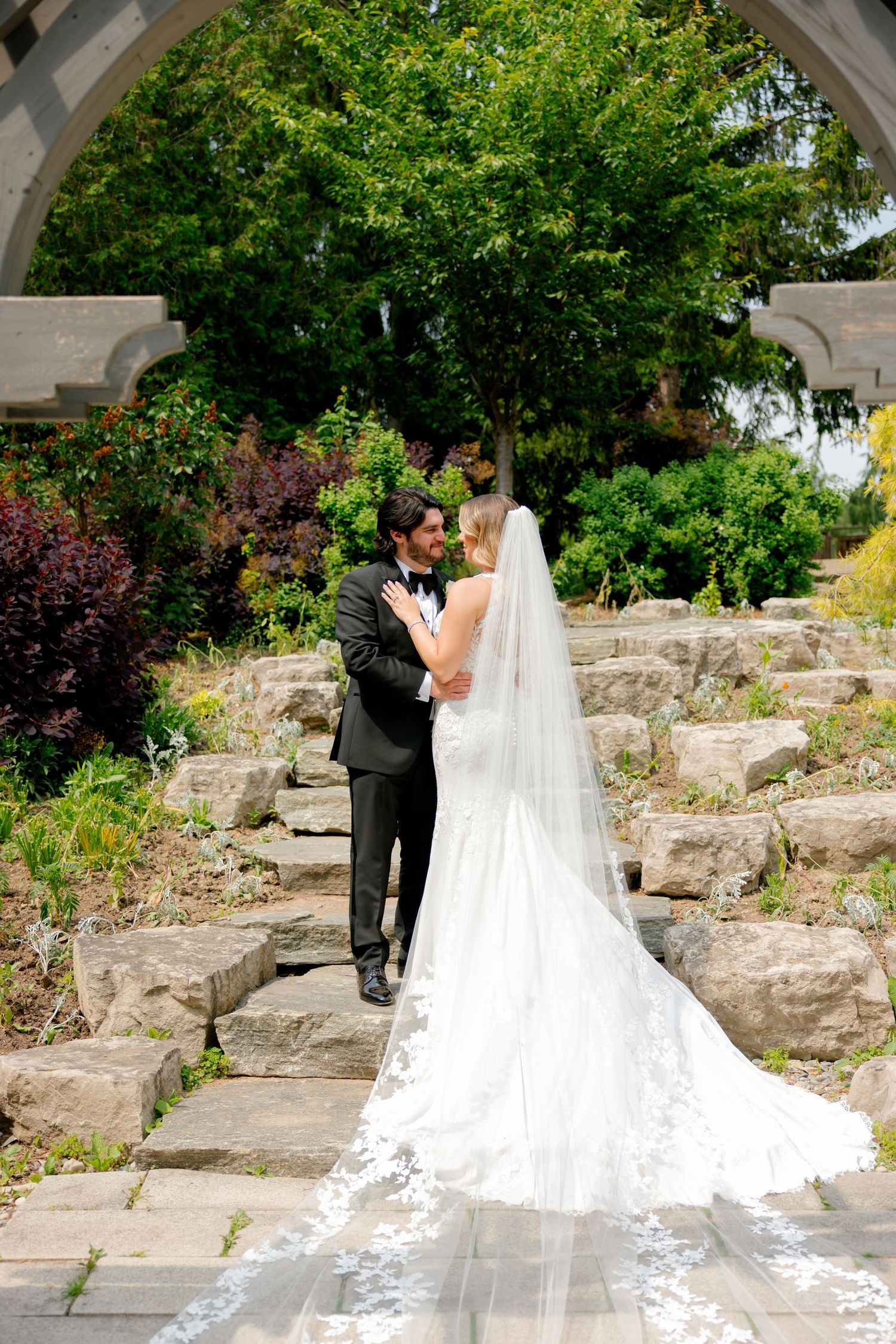 Romantic wedding portrait under garden archway with long lace veil cascading down stone staircase.