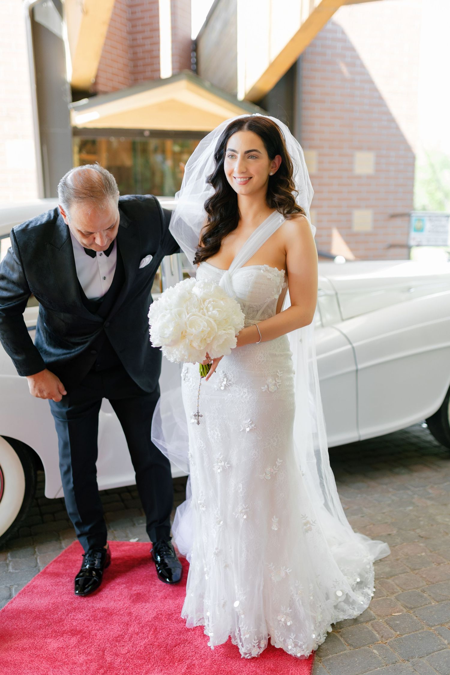 A beautiful bride in a white lace gown holds a bouquet next to a classic white car on a red carpet.