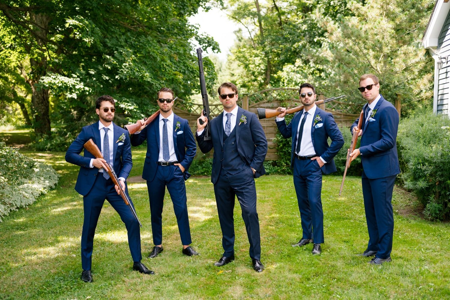 Groomsmen in navy suits strike playful poses on a green lawn during a wedding photoshoot.
