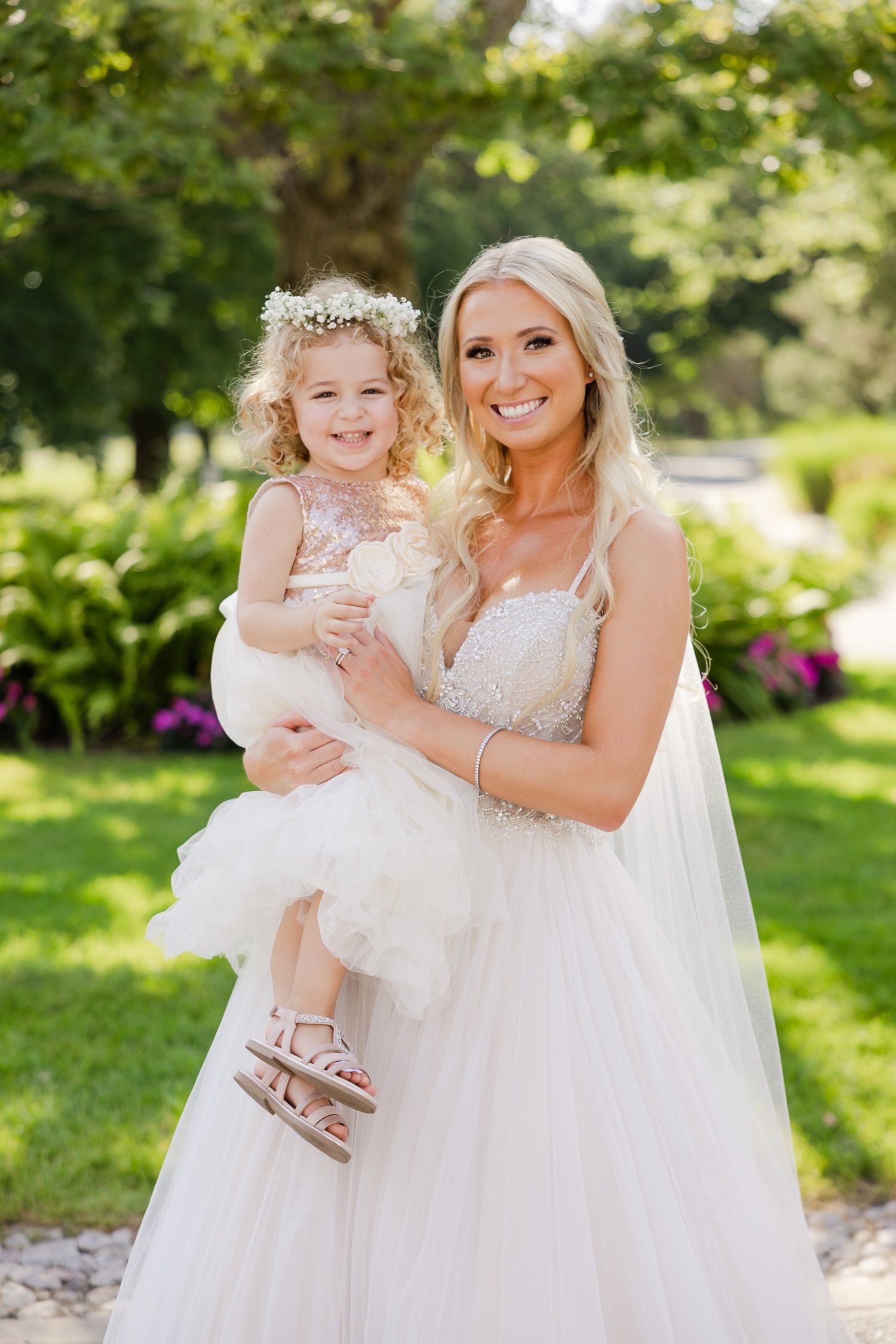 A bride in a white lace gown holds a young girl in a matching tulle dress in a garden setting.
