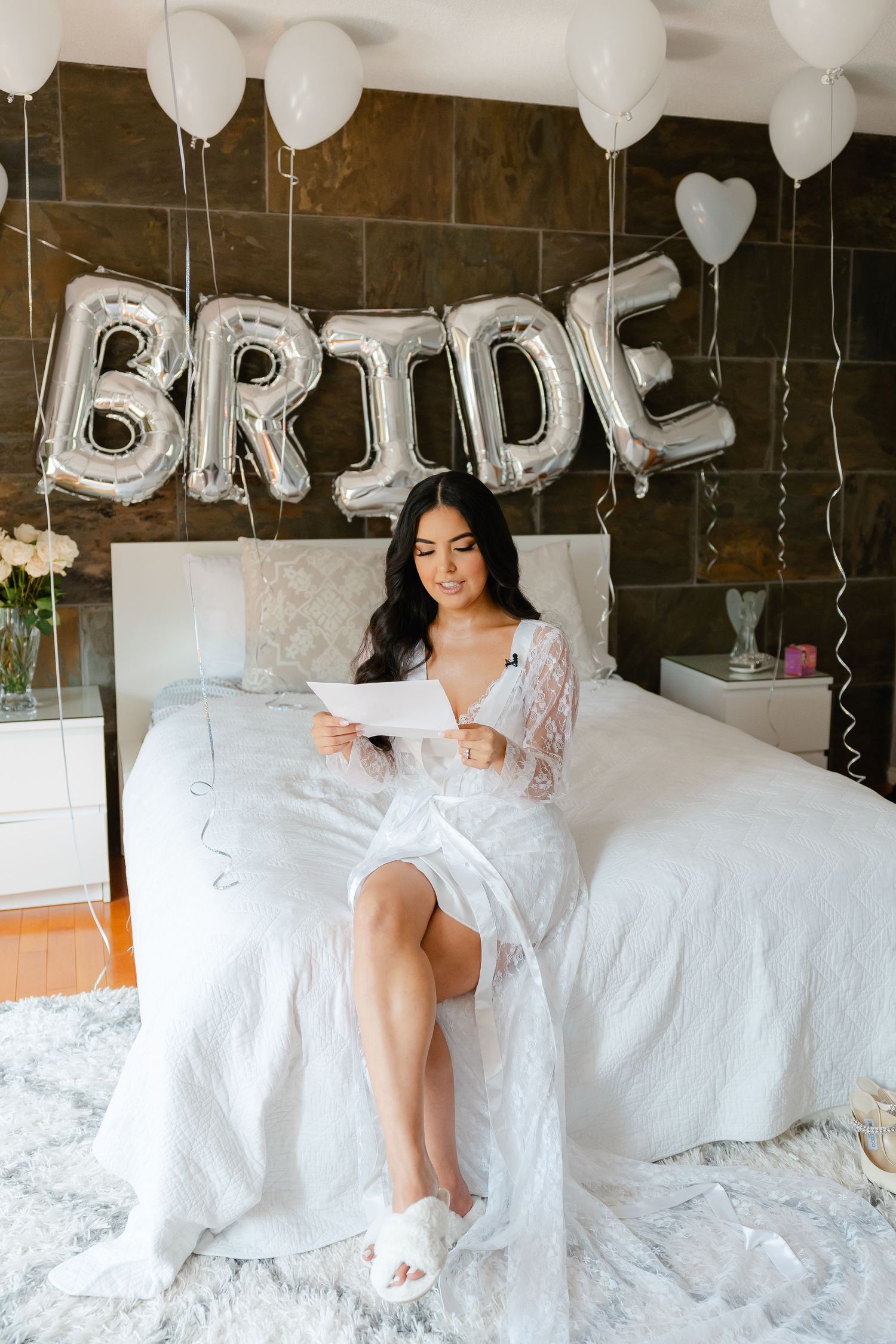Person in white robe sits on bed under silver BRIDE balloon letters and white decor.