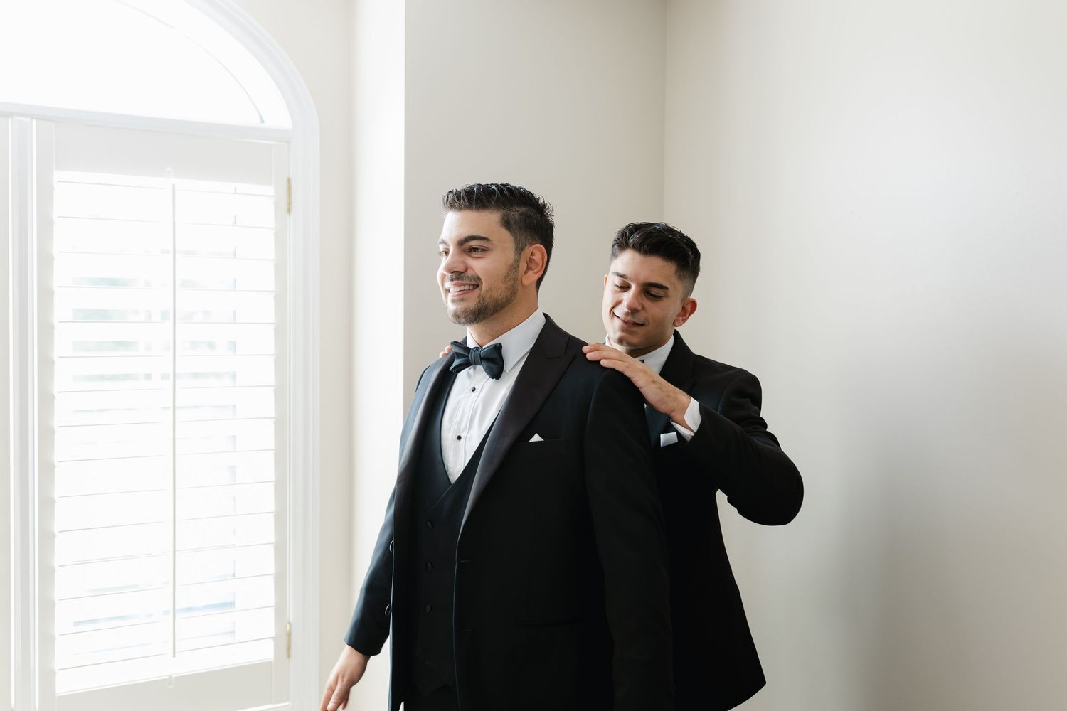 Two people in matching tuxedos help each other prepare for a formal event.