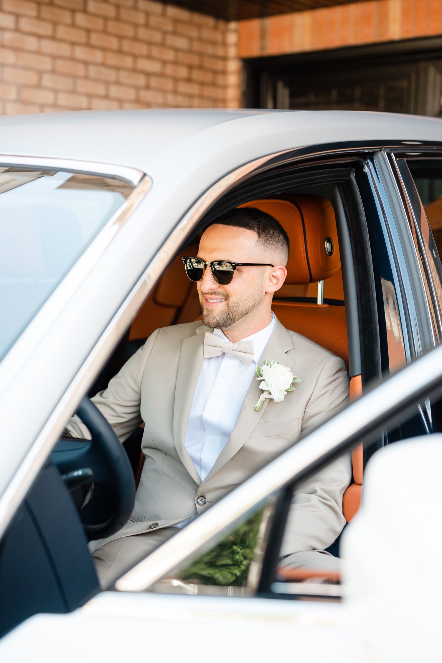 Groom in beige suit and bowtie smiles while sitting in luxury car with tan leather interior.