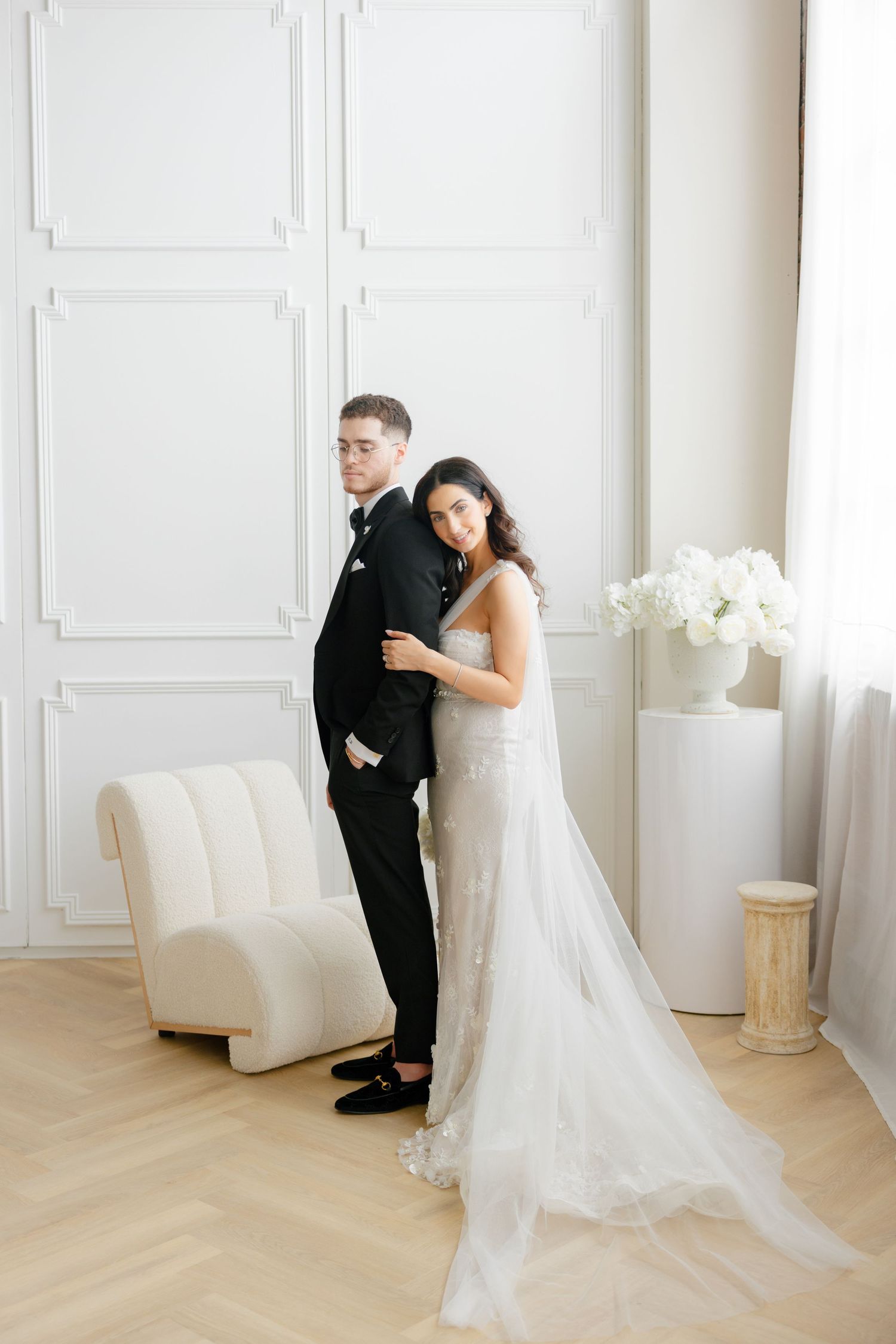 An elegant wedding pose with the couple standing before white paneled walls and soft window light.