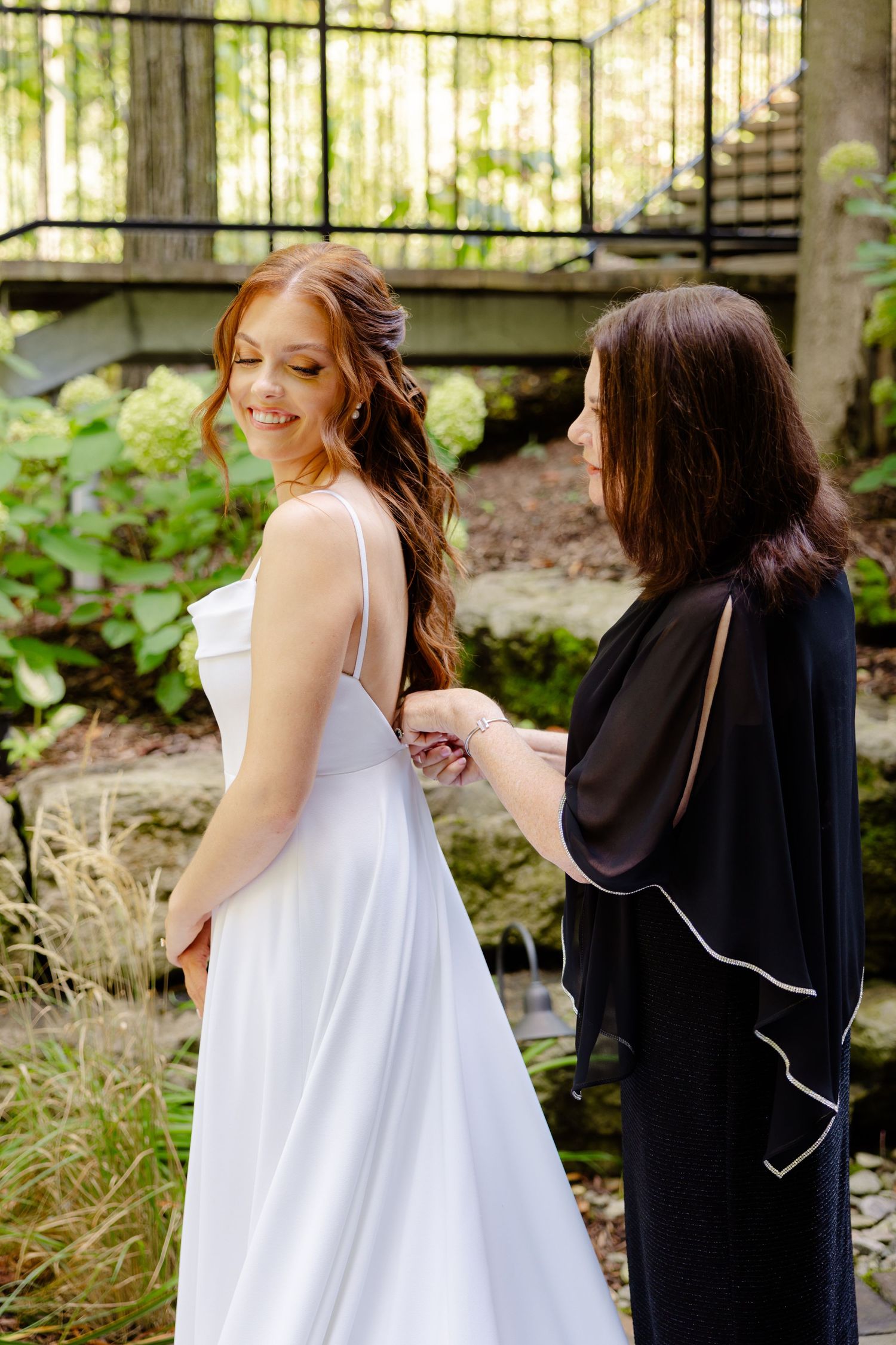 Candid moment of bride getting dressed with assistant in garden setting.