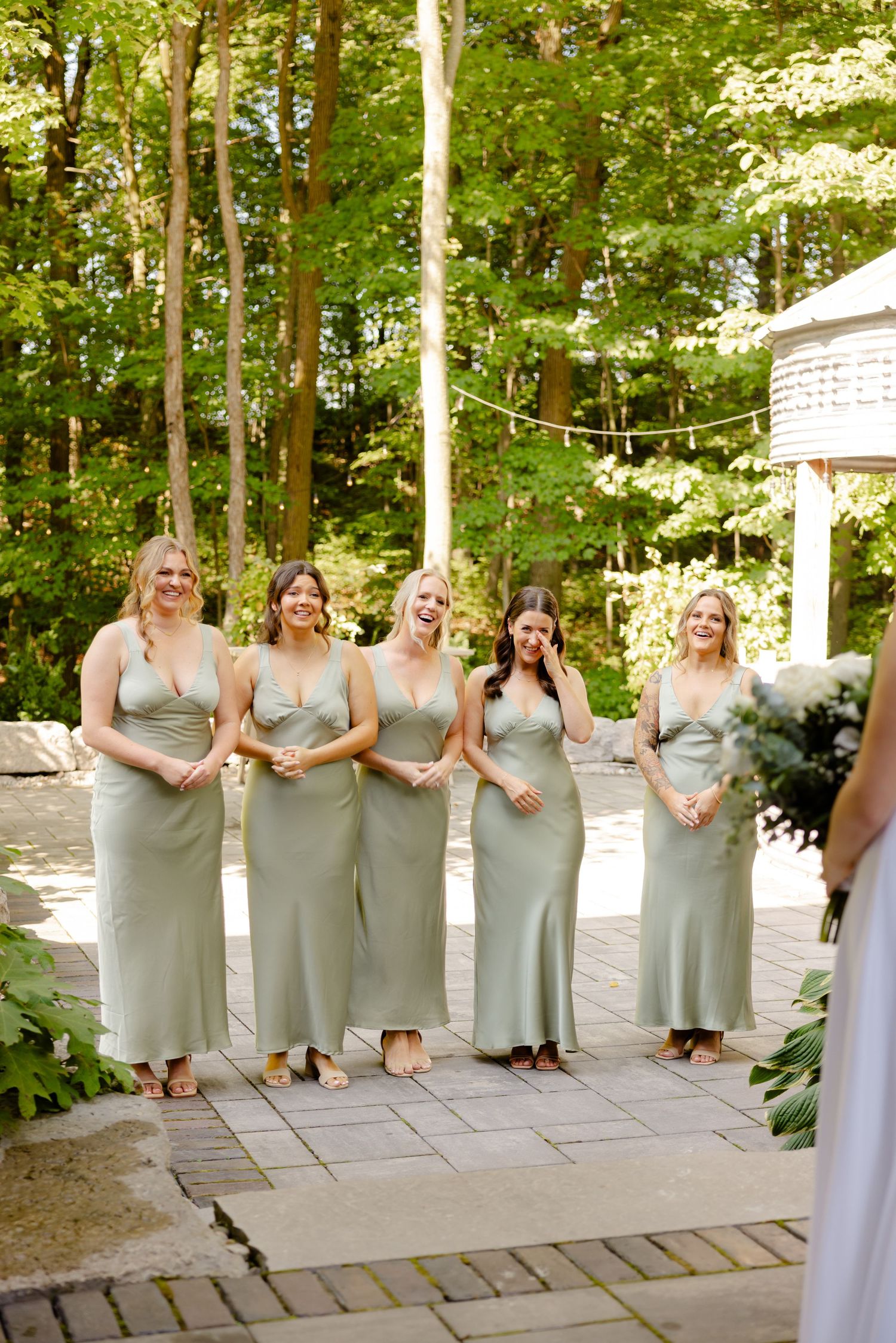 Wedding party in matching sage dresses poses on outdoor stone patio with greenery backdrop.