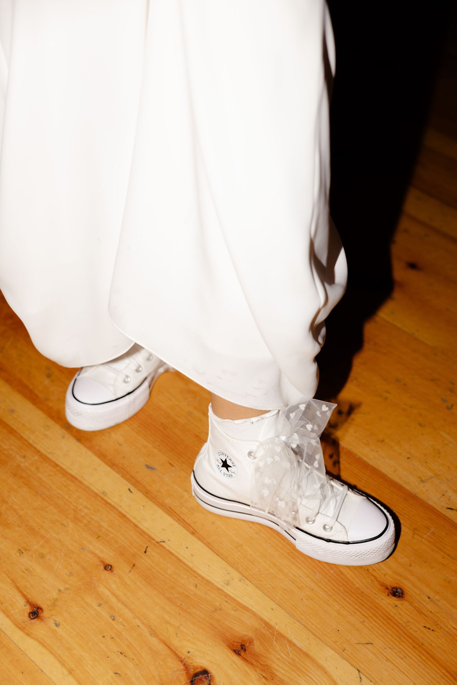 White Converse sneakers peek out under a flowing wedding dress on a wooden floor.