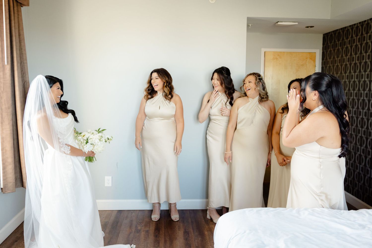 Bride in white dress shares moment with bridesmaids wearing cream colored dresses in hotel room.