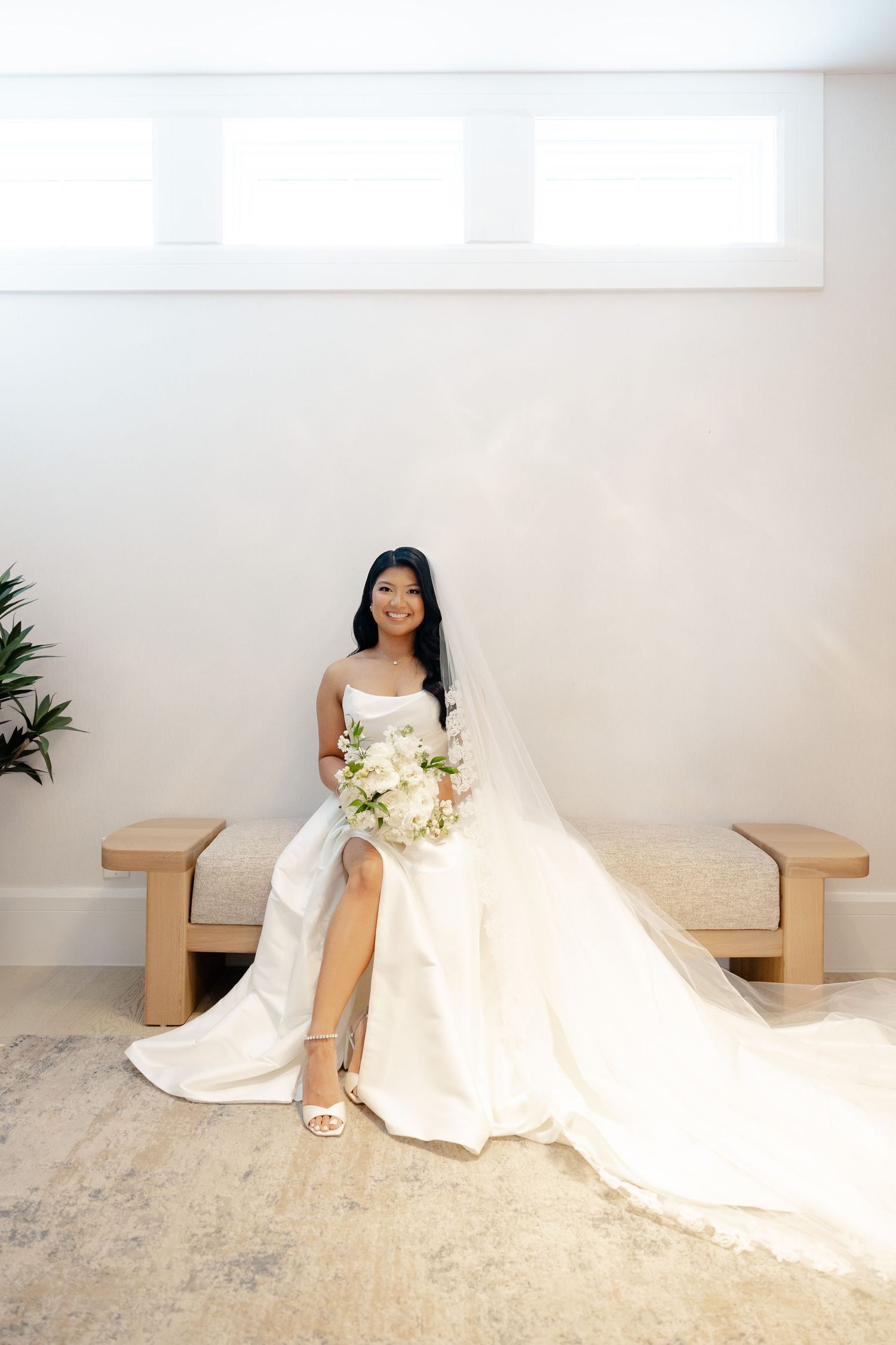 Bride in elegant white gown sits on wooden bench holding bouquet in minimalist room with long flowing train.