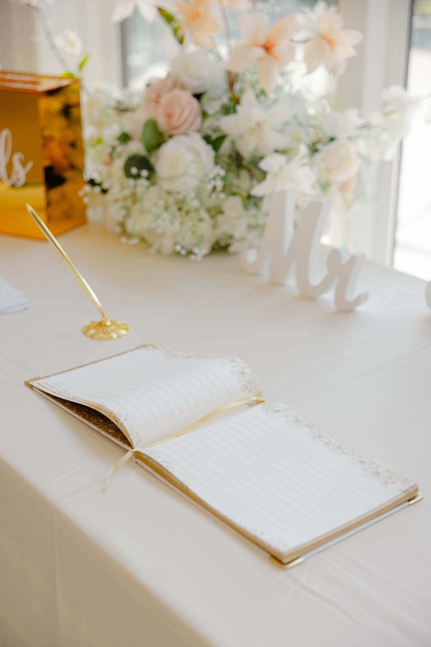 Open guestbook with gold pen on white table beside delicate floral arrangements.
