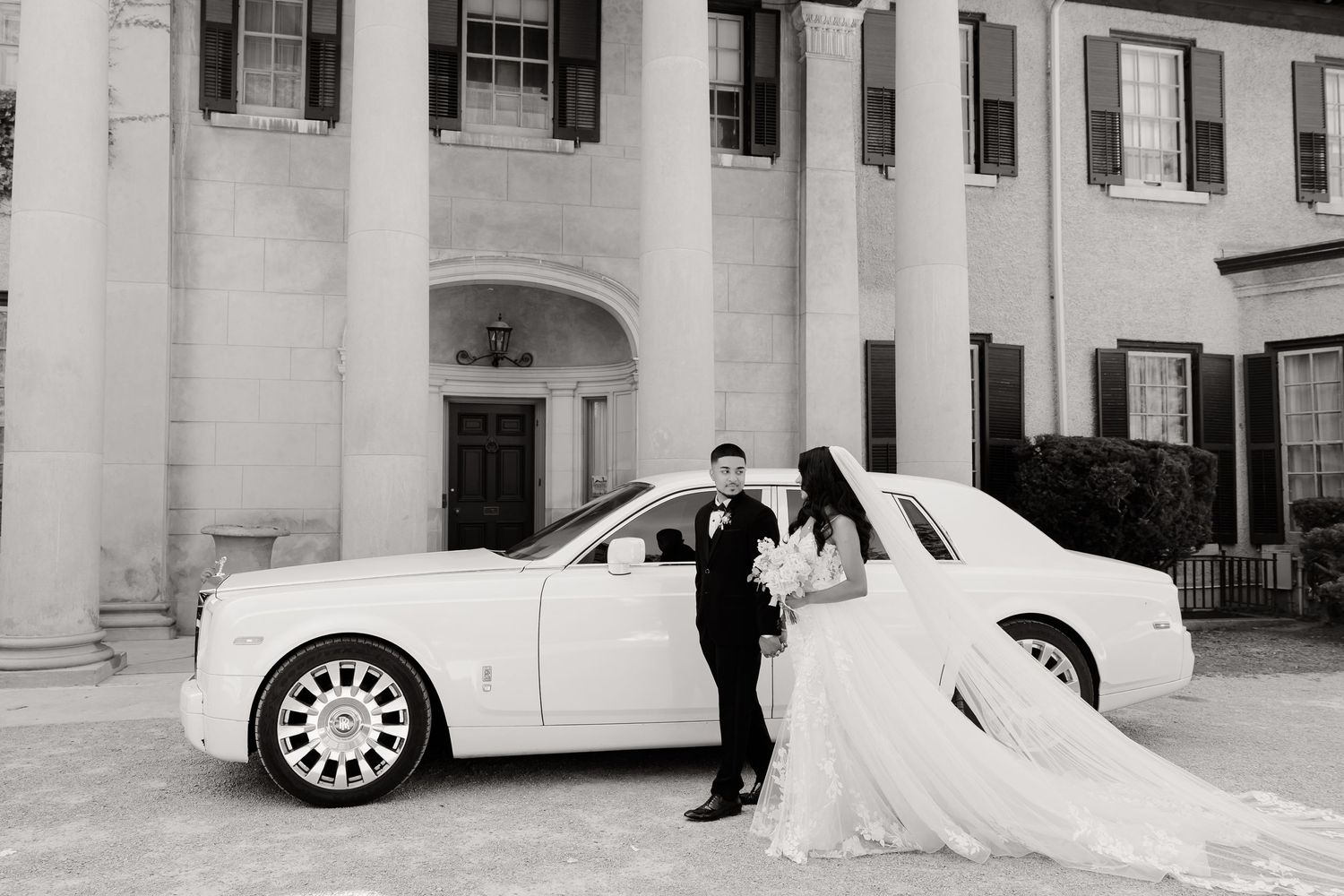 A luxury white car with a couple in wedding attire standing beside it in front of a grand white building.