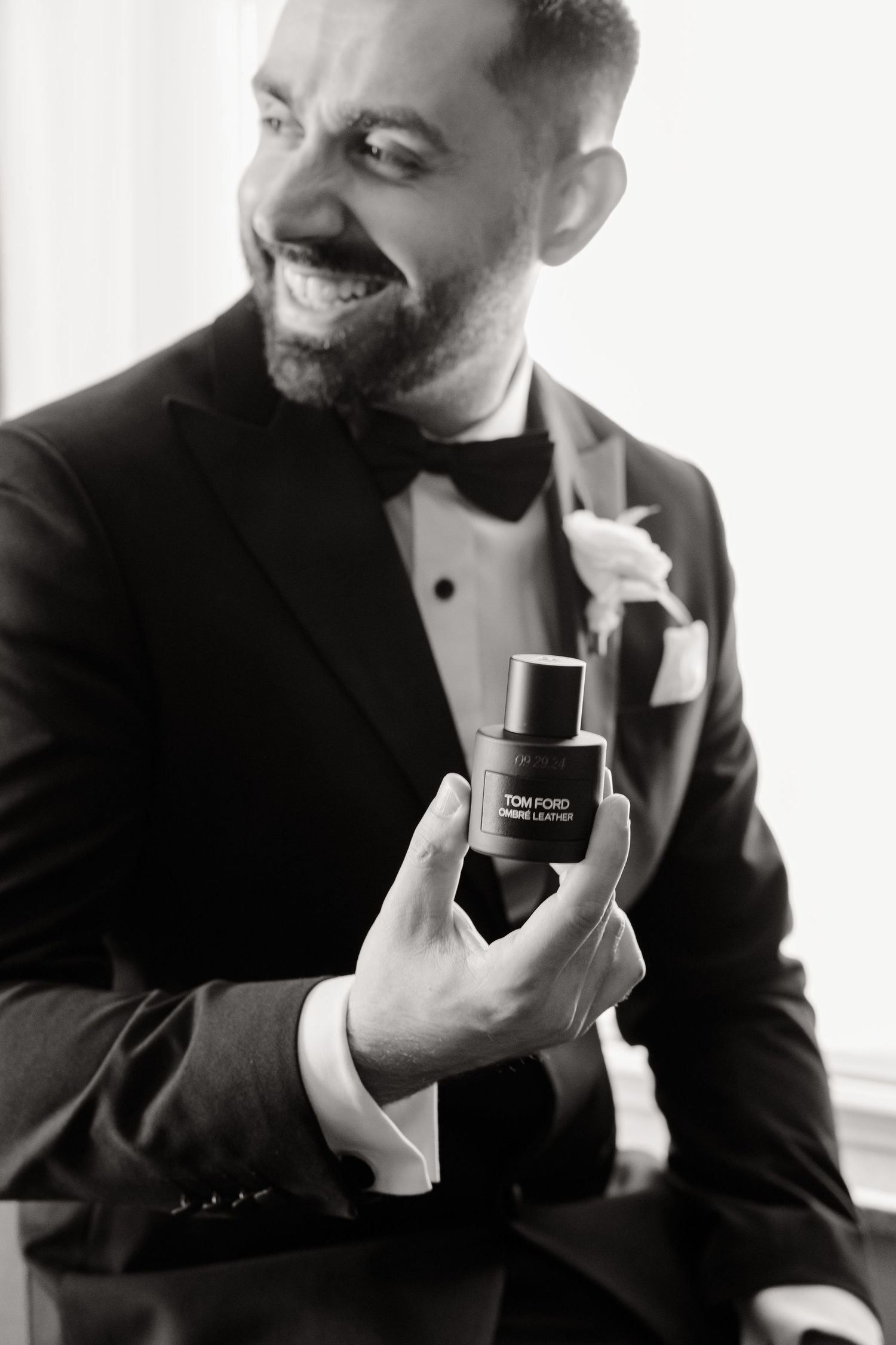 Black and white photo of groom holding cologne bottle while smiling.
