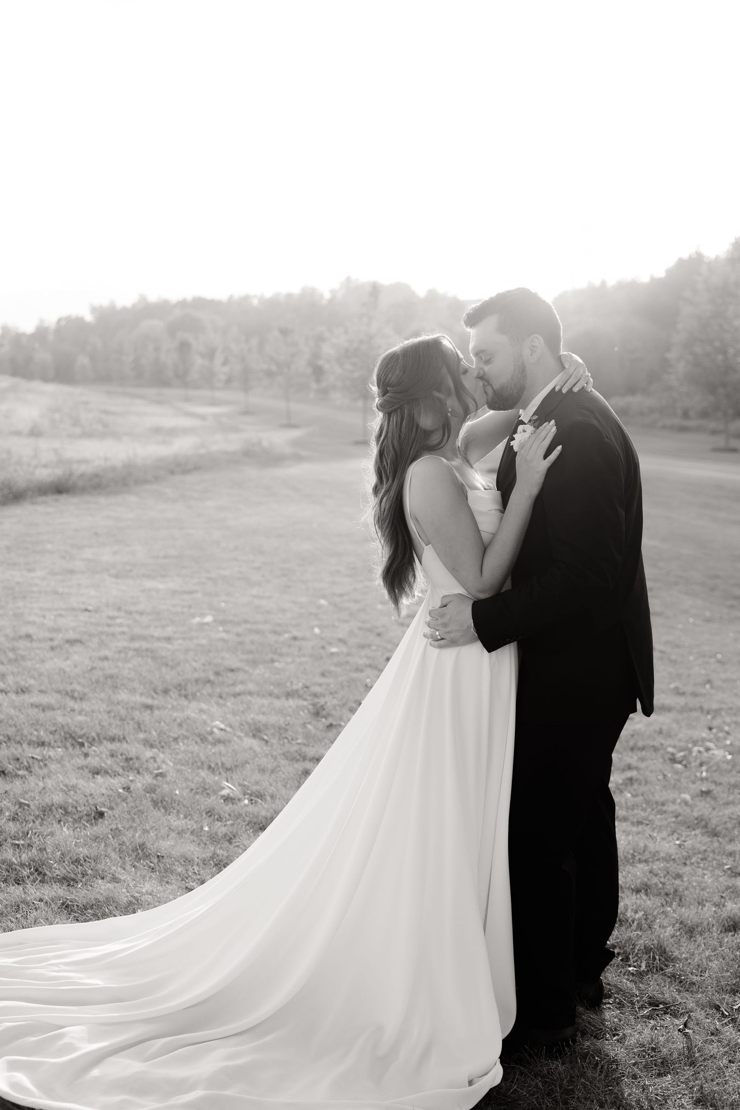 Dramatic black and white wedding photo shows couple embracing in field with flowing white dress train.
