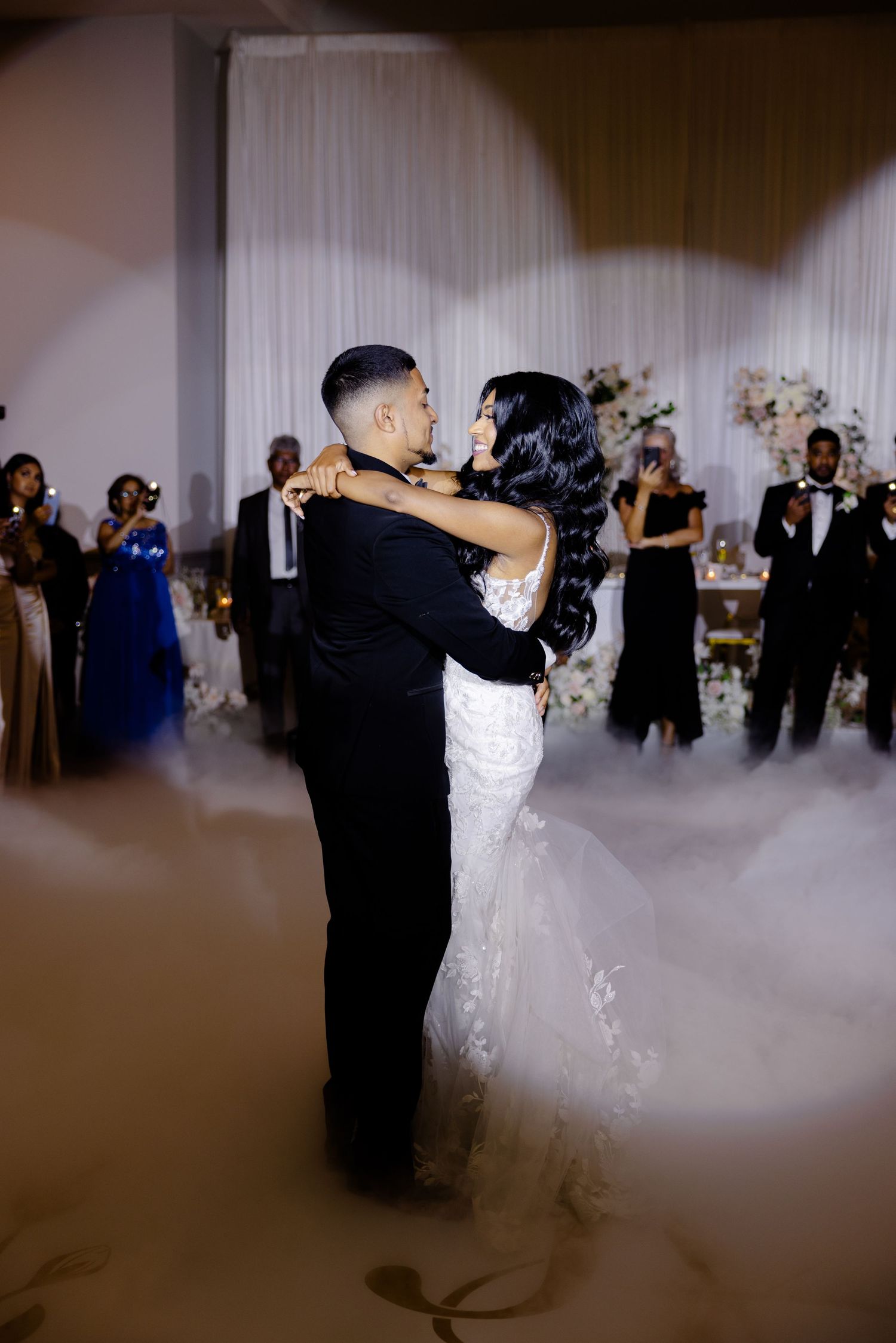 A couple embraces during their first dance with guests and floral arrangements in the background.