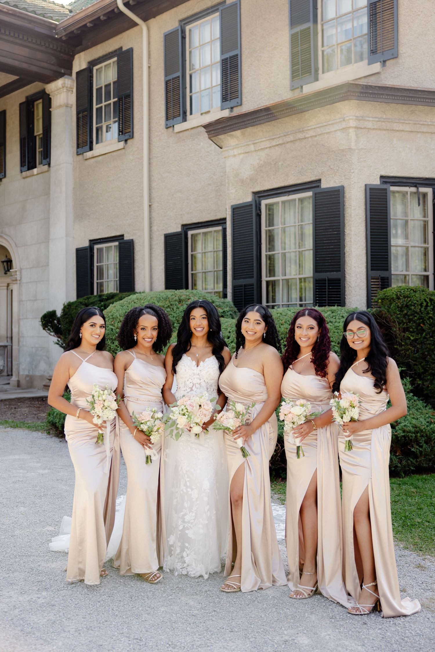 Wedding party in matching champagne dresses pose together outside historic mansion.