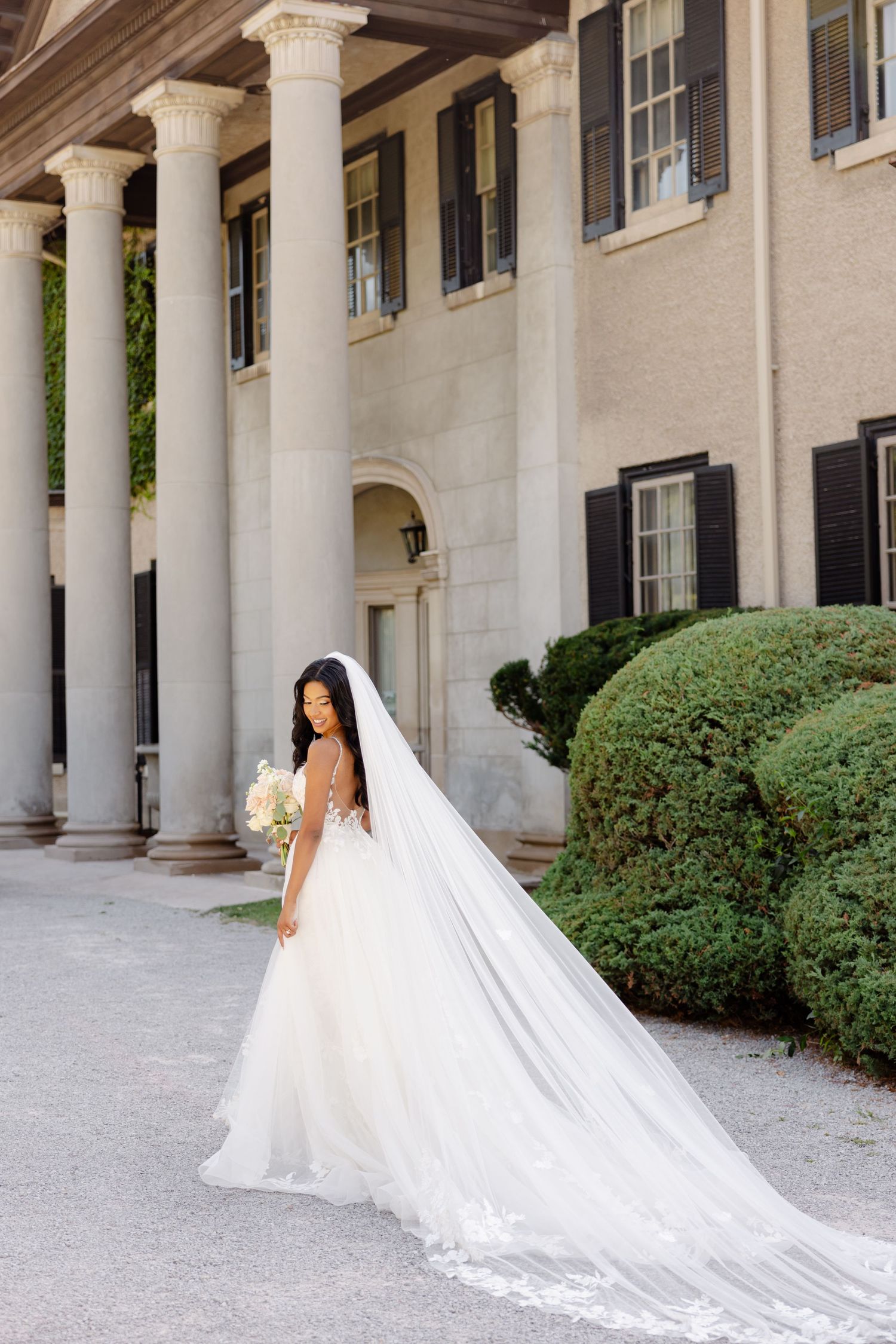 A wedding photo capturing a bride in a flowing cathedral veil against a historic mansion backdrop.