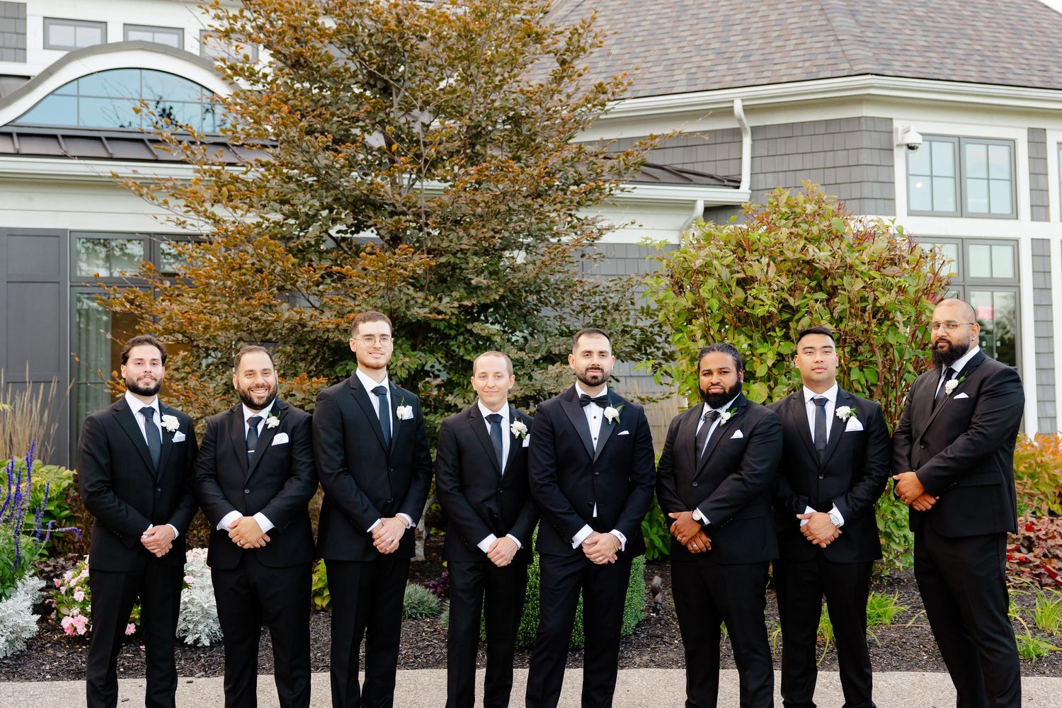 Groomsmen in black suits pose for formal photo outside gray wedding venue with fall foliage.