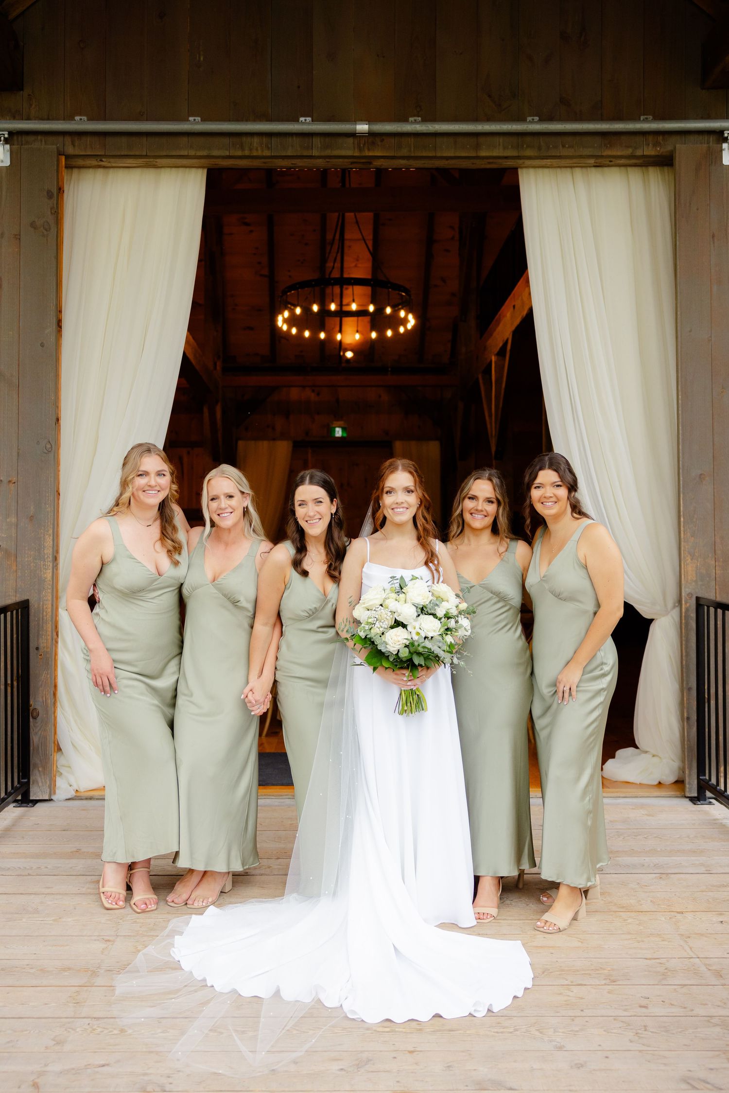 Bridal party in sage green dresses poses with bride in white gown at rustic venue entrance.