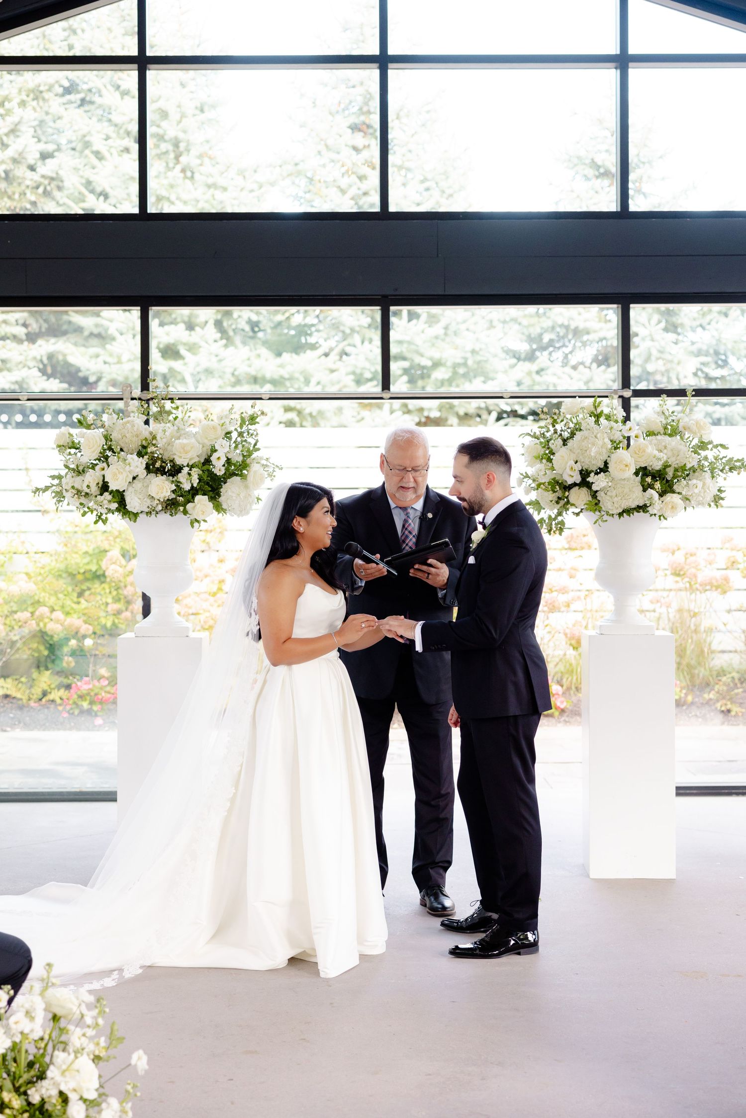 A wedding ceremony with white floral pedestals and large windows creating a bright, airy atmosphere.
