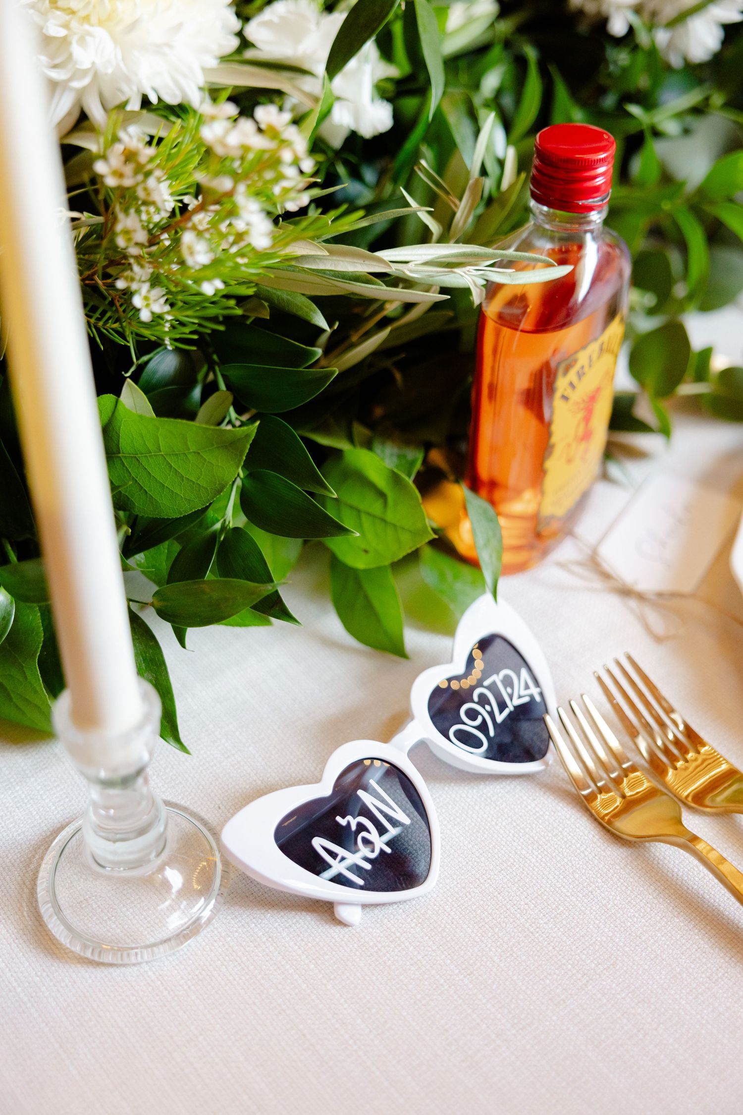 Wedding reception table setting with heart-shaped signs, candles, greenery and a bottle of spirits.