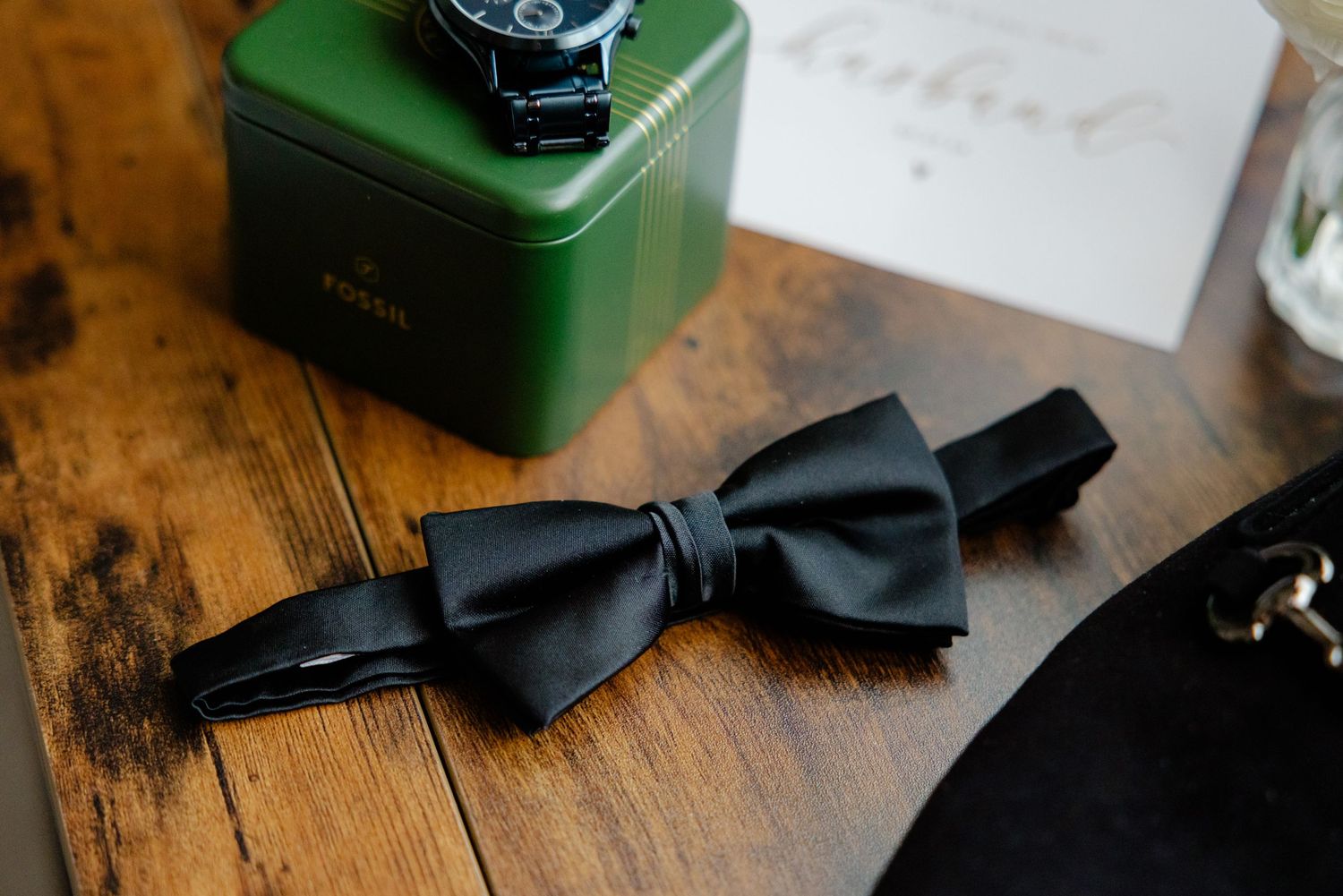 Black bow tie laid flat on wooden surface near wedding accessories.