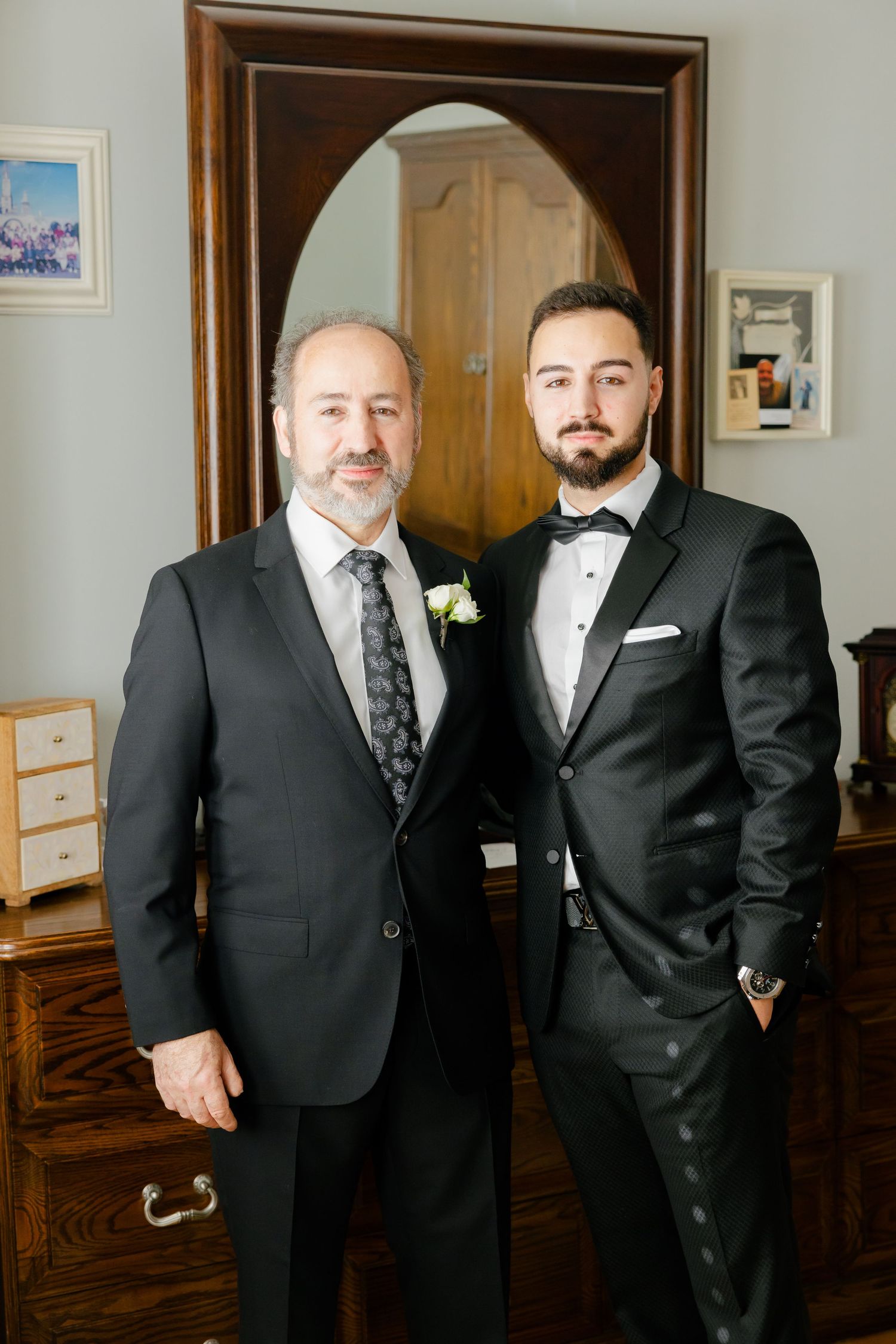 Two people in formal suits standing together in front of a framed mirror and wooden dresser.