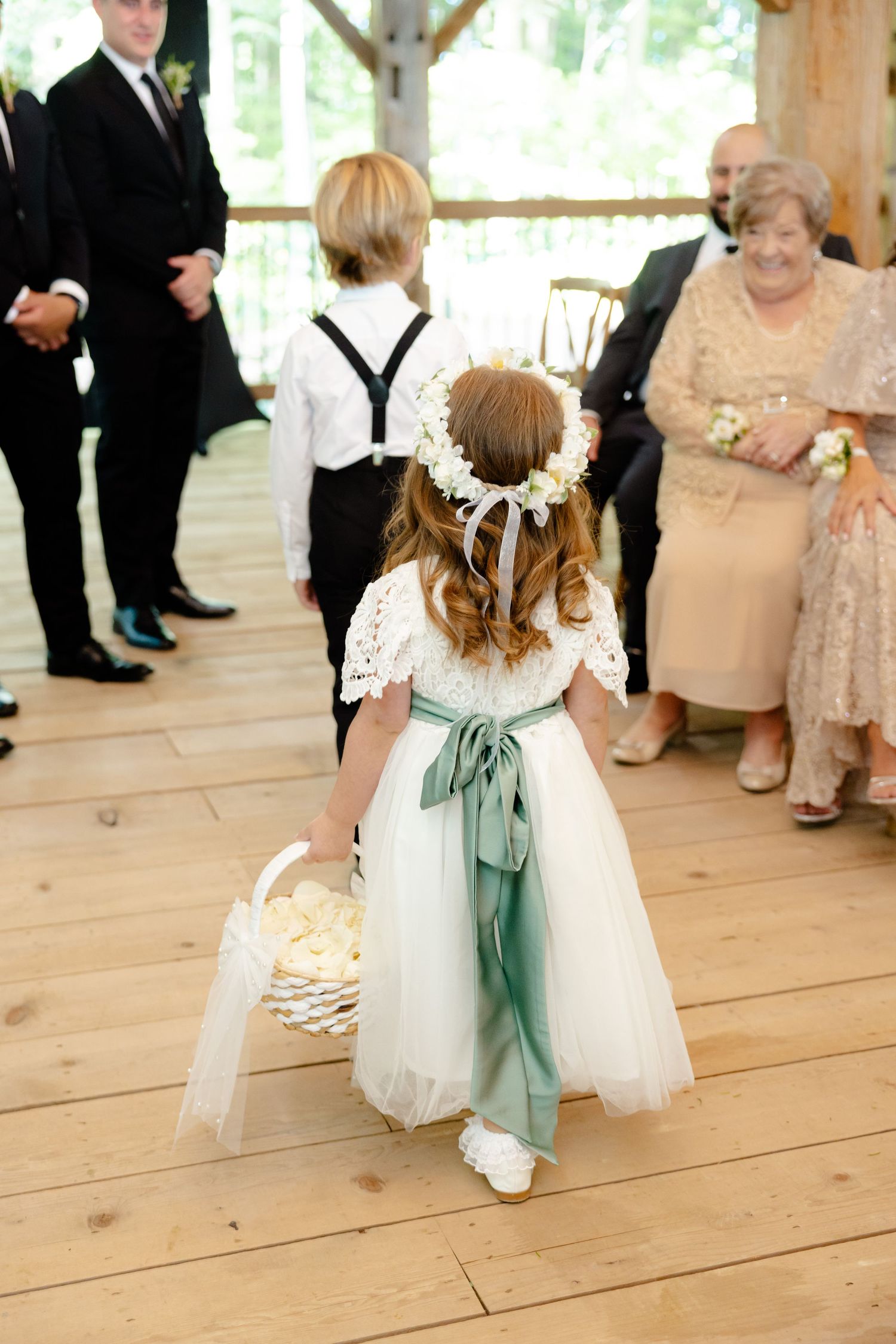 A small flower girl in a white dress with green sash carries a basket down a wooden aisle.