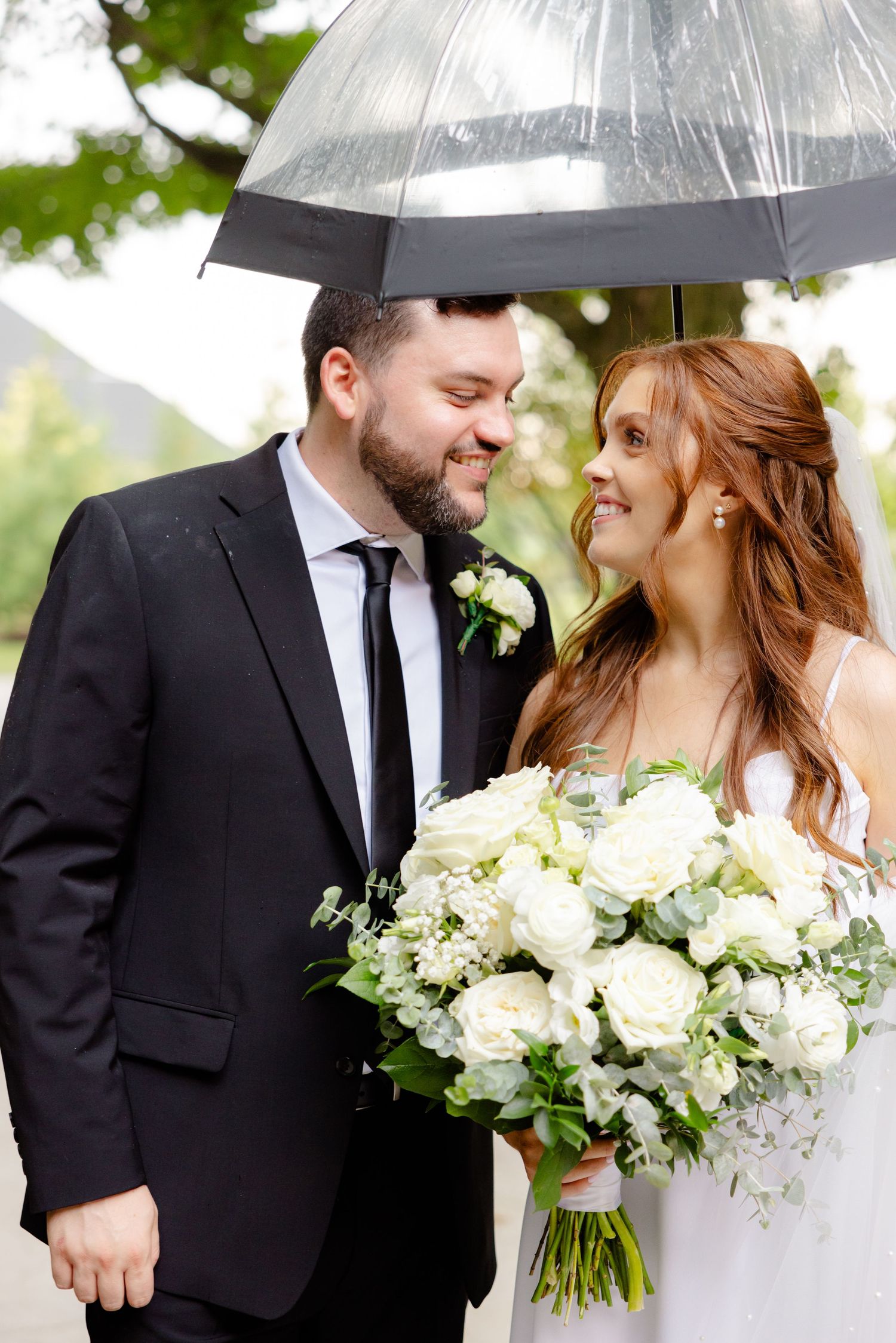 Couple smiles at each other under clear umbrella while holding elegant white bouquet.