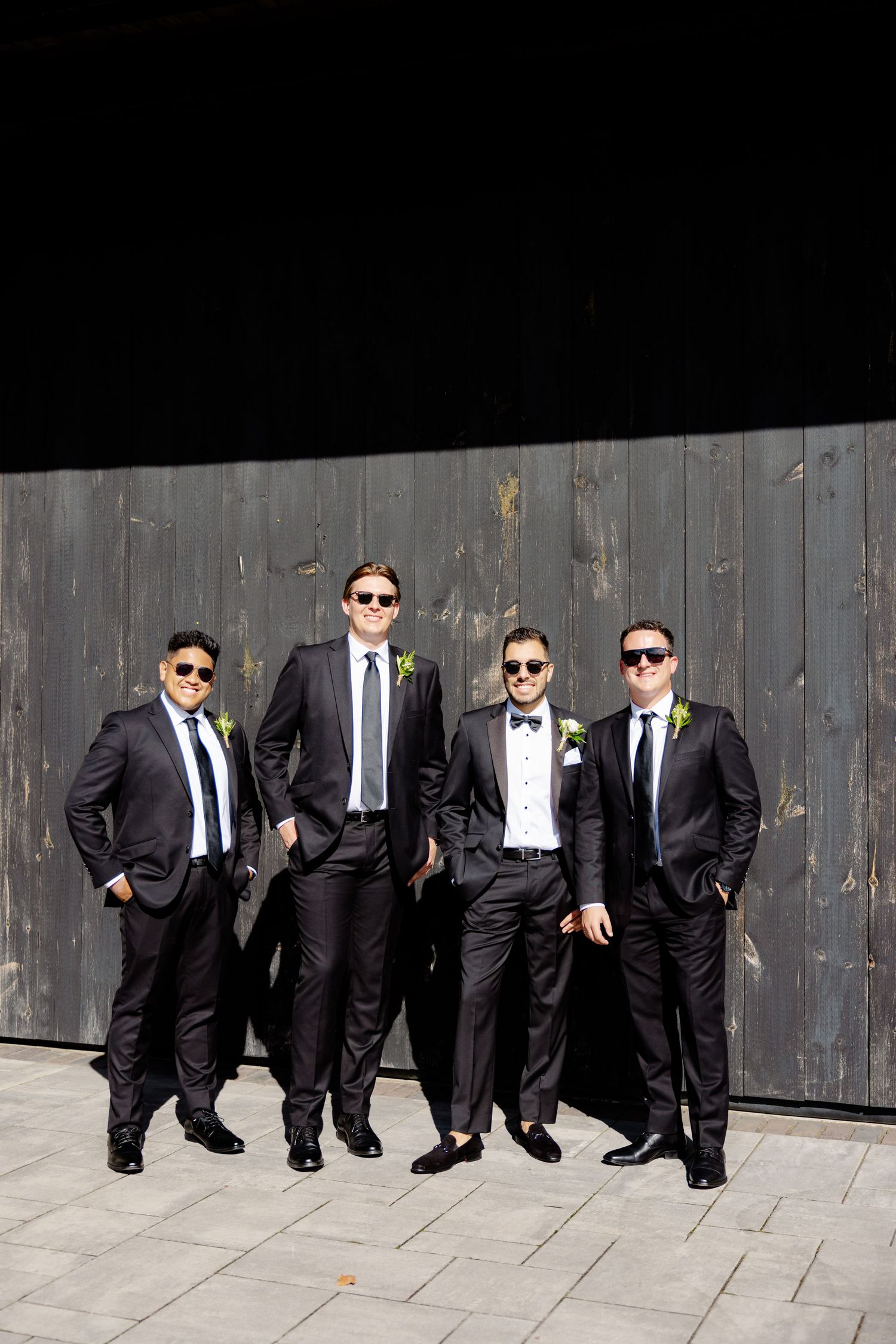 Groomsmen in dark suits and sunglasses pose against rustic wooden wall.