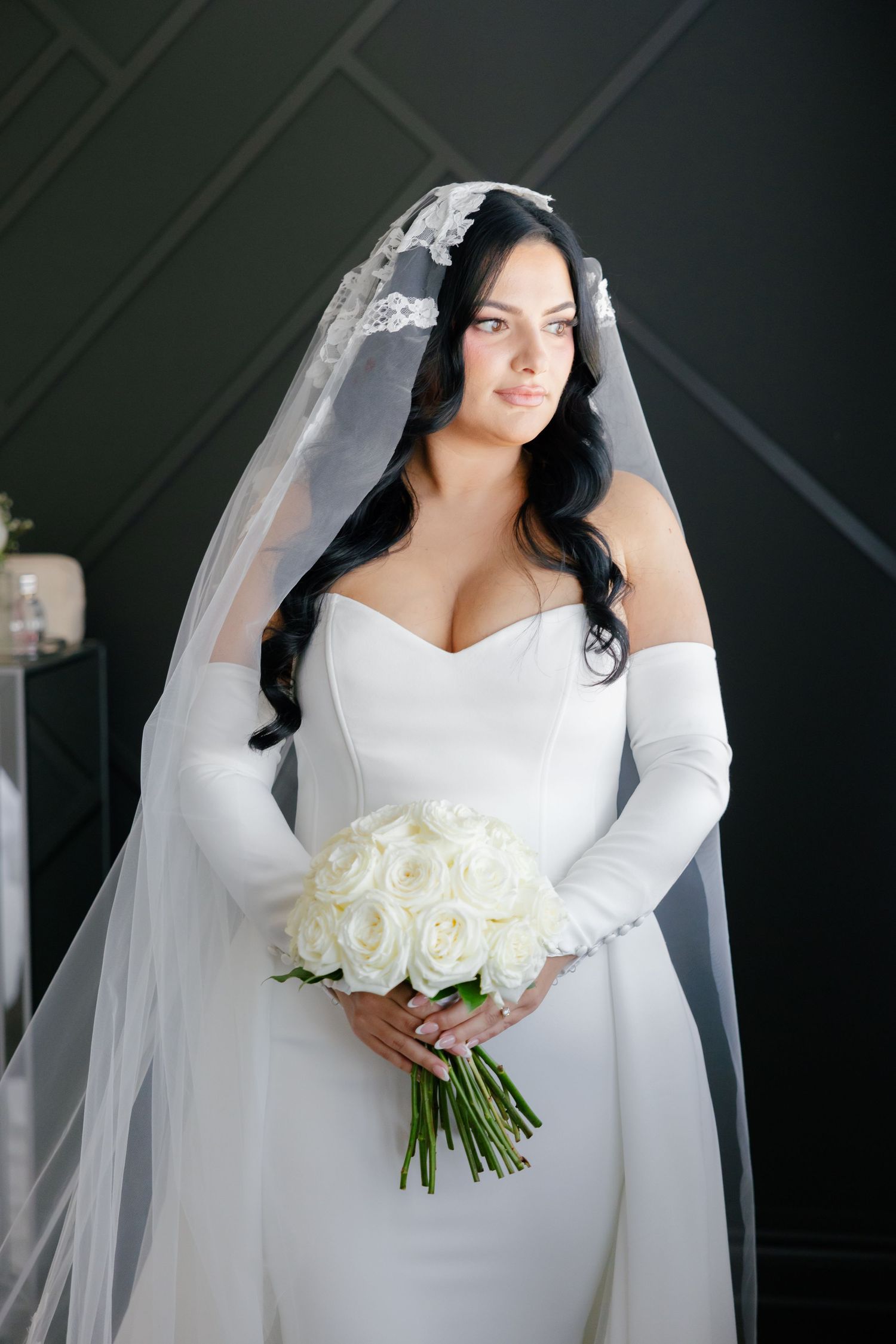Elegant bride in white fitted gown holds white rose bouquet against dark wall.