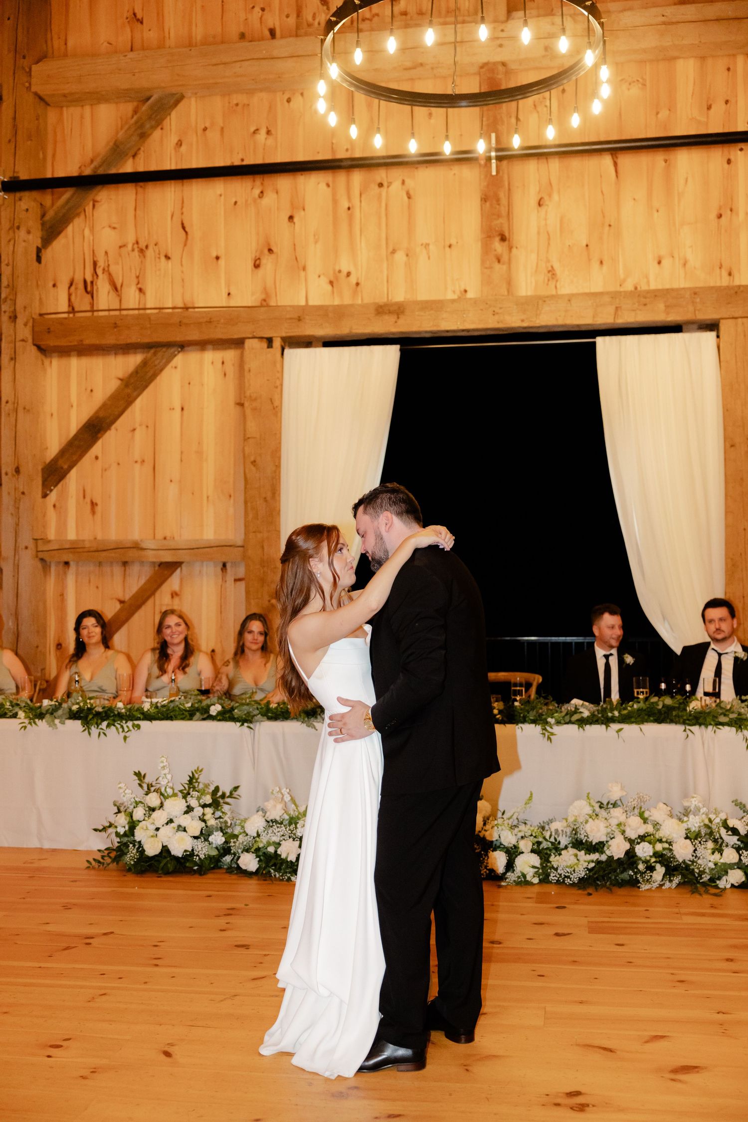 First dance beneath wooden beams and chandelier with wedding party seated at decorated head table.