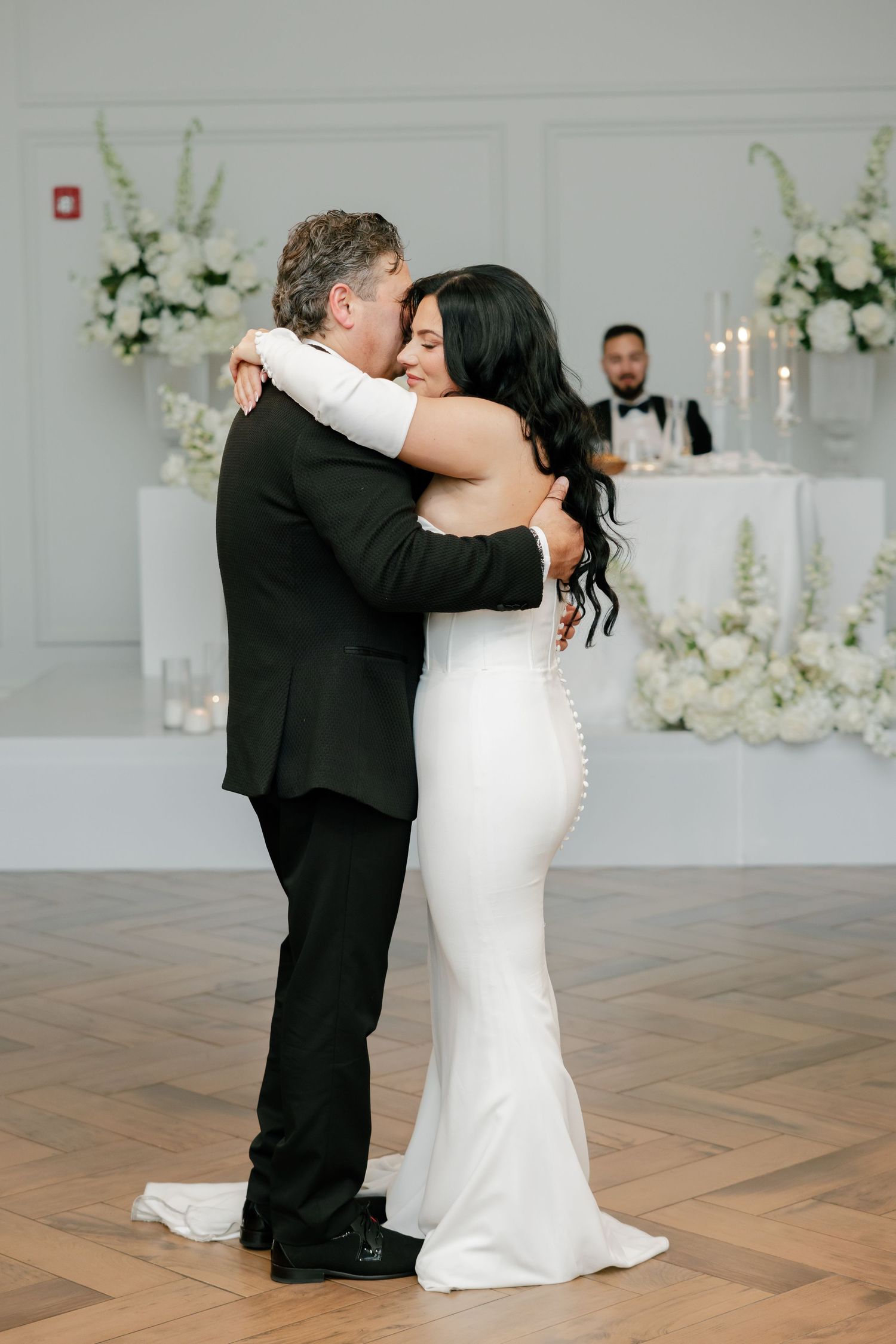 Wedding couple embraces during emotional dance moment surrounded by white floral arrangements and candlelight.