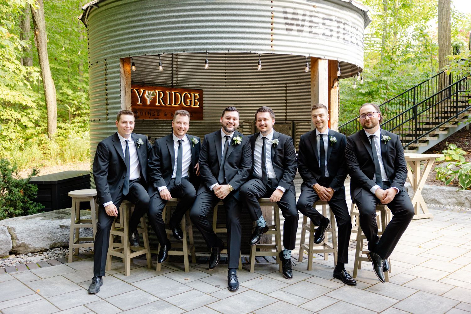 Wedding party members in dark suits pose casually on bar stools at outdoor venue.
