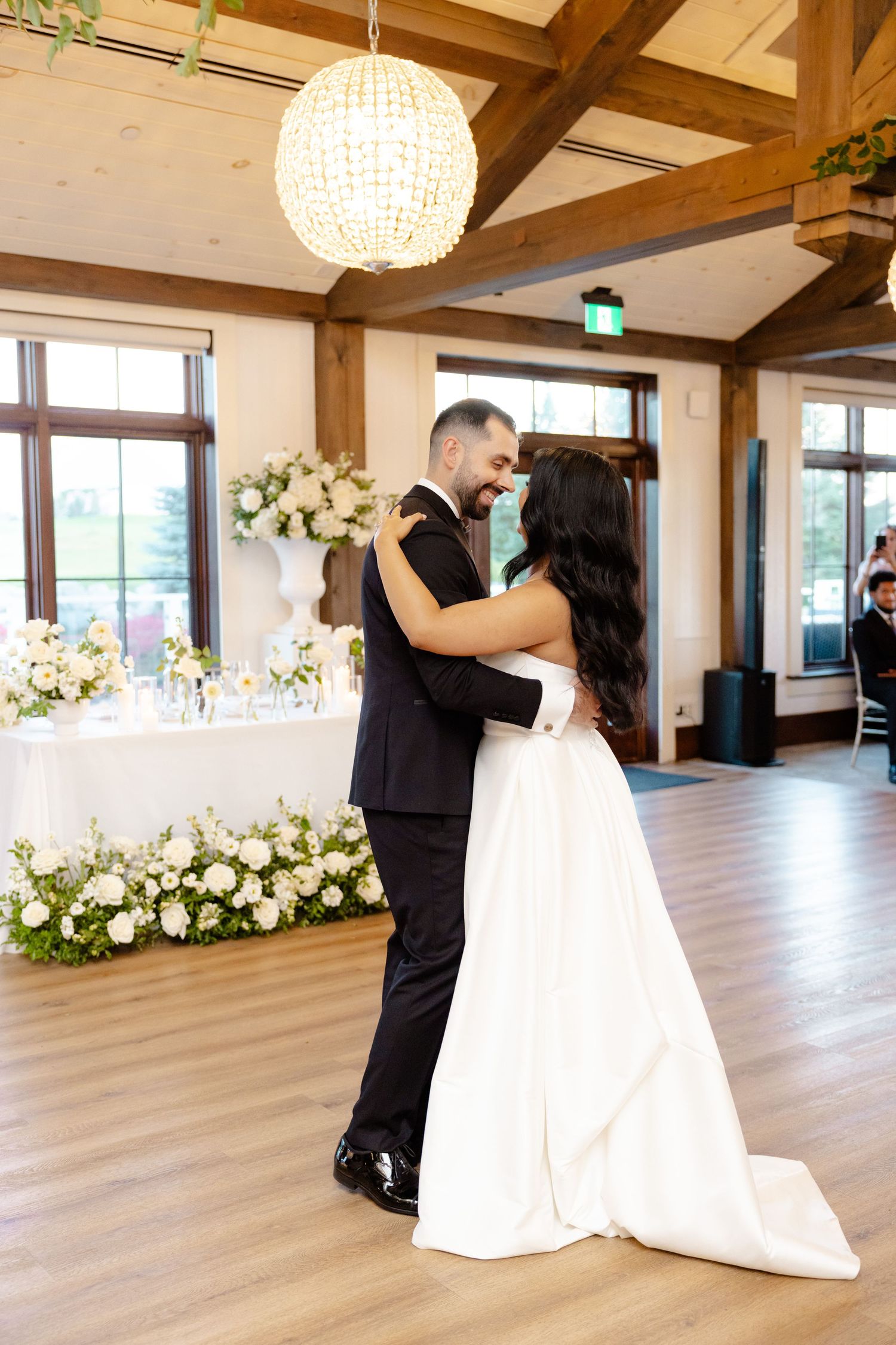 A couple shares their first dance in an elegantly decorated room with white floral arrangements and a crystal chandelier.