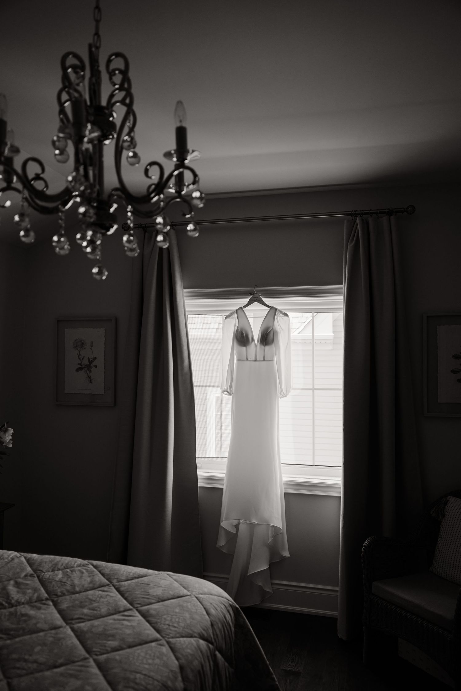 Elegant wedding dress hanging in window with chandelier in moody black and white photograph.