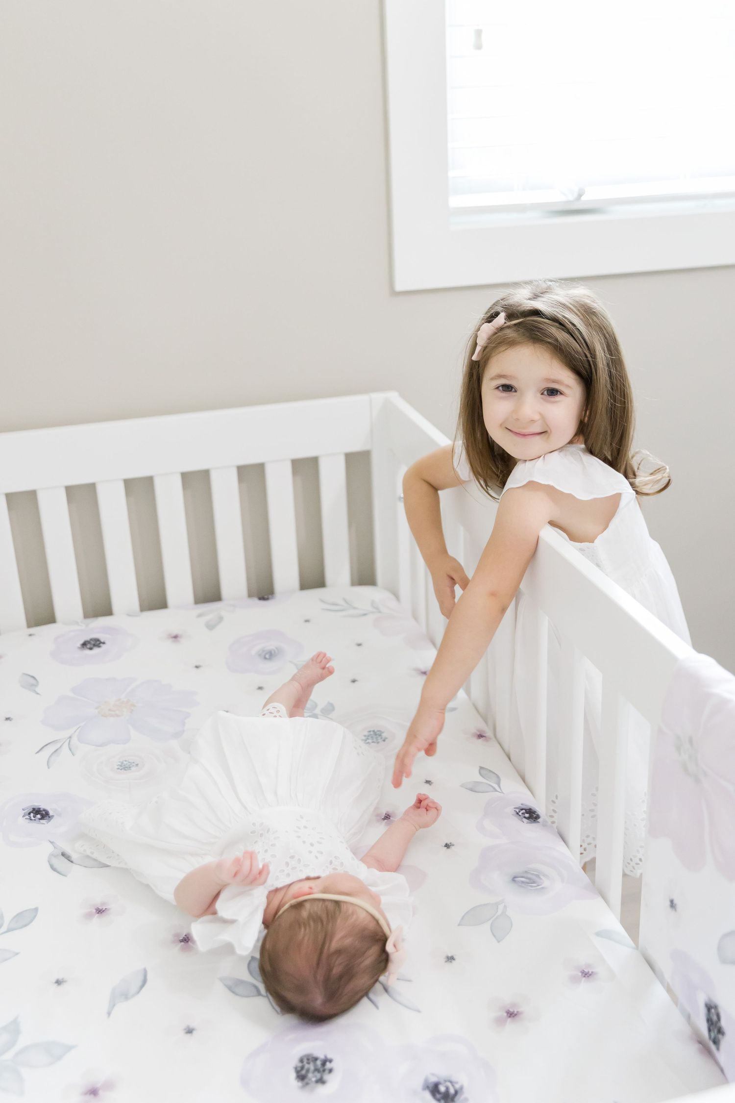 Child smiles while standing at crib containing sleeping newborn on floral bedding.