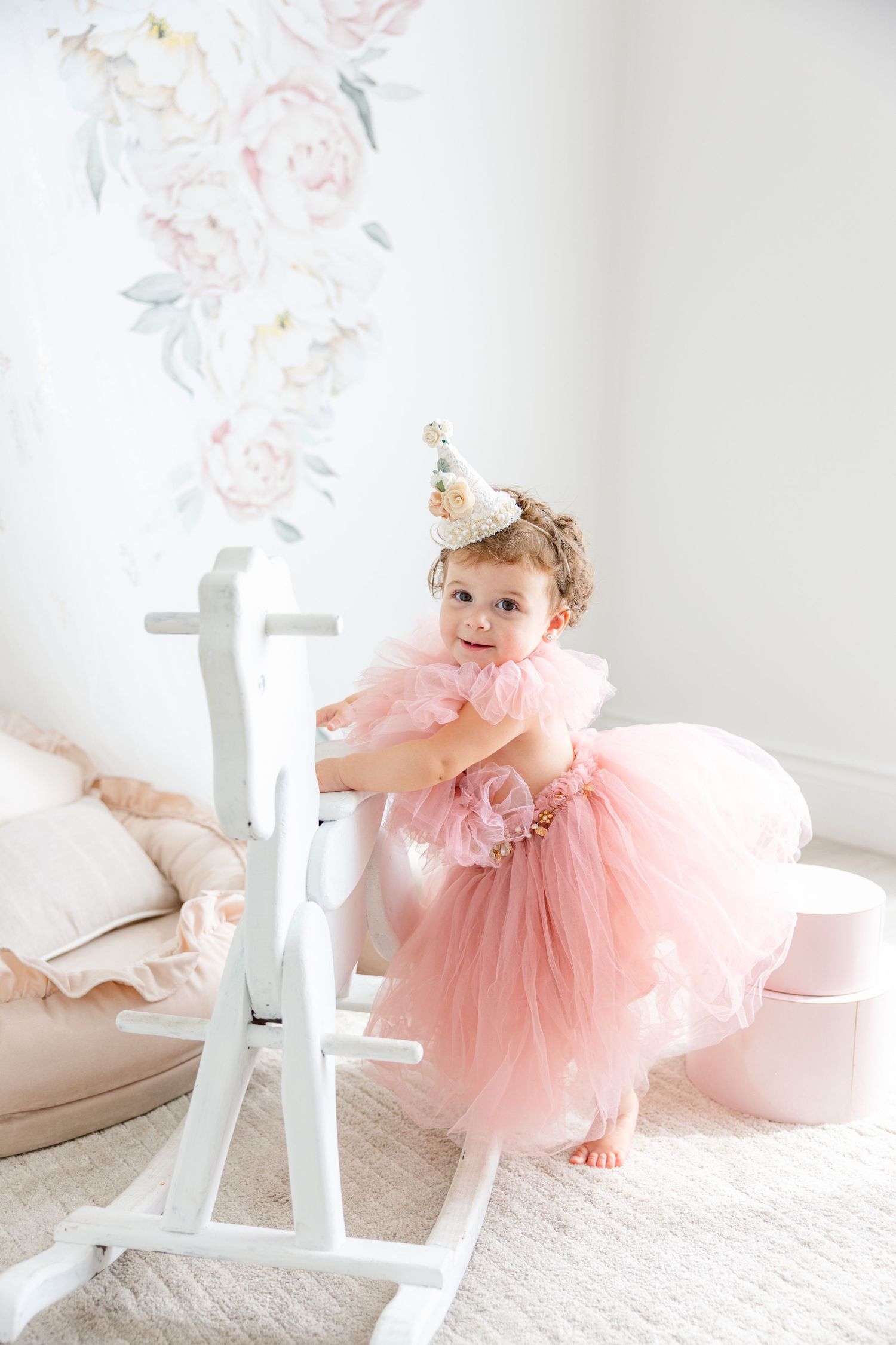 Whimsical nursery scene with white rocking horse and pink tulle dress against floral wallpaper.