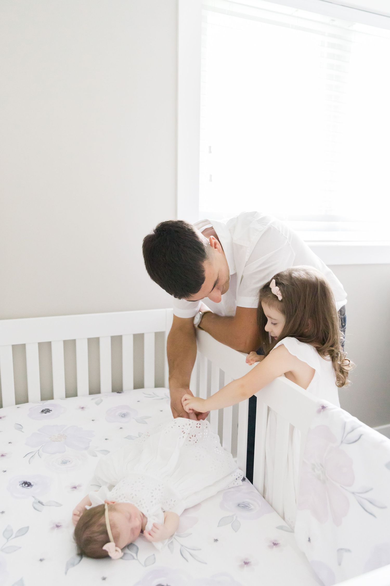 Parents looking lovingly at their sleeping baby in white crib by window.