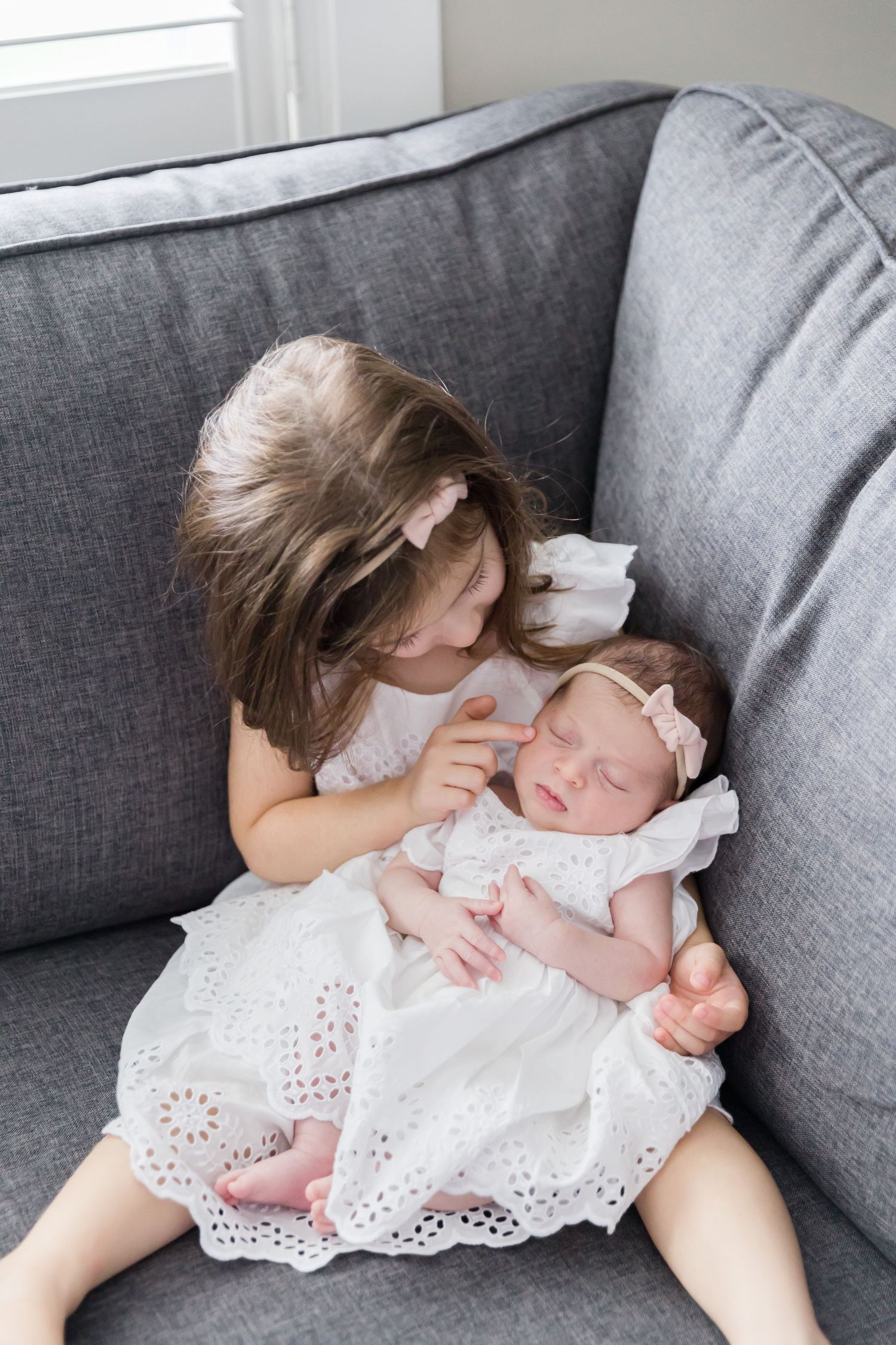 Two children in white eyelet dresses sitting together on a grey couch showing sisterly affection.