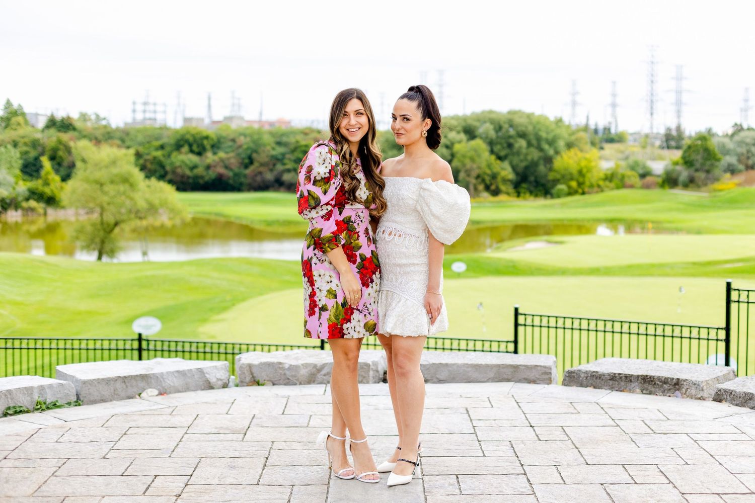 Two friends in floral and white dresses pose together on stone patio with golf course view.