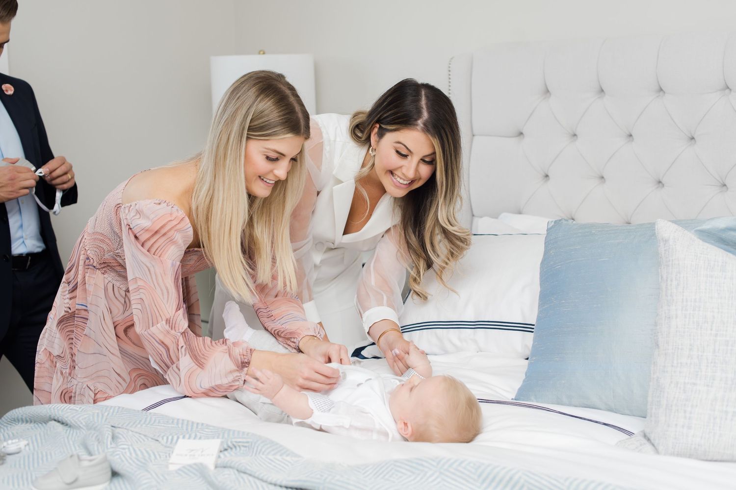 Women in pink and white dresses attending to infant on bed.