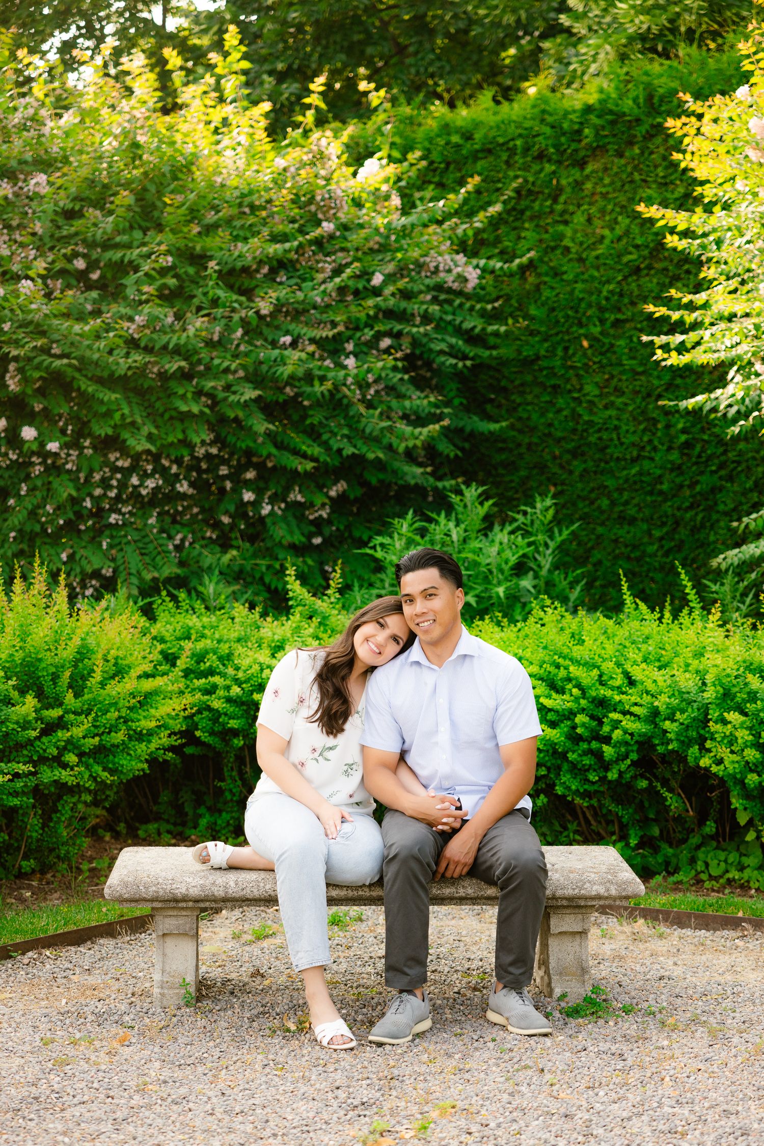 A couple sits together on a stone bench surrounded by lush green garden foliage on a sunny day.