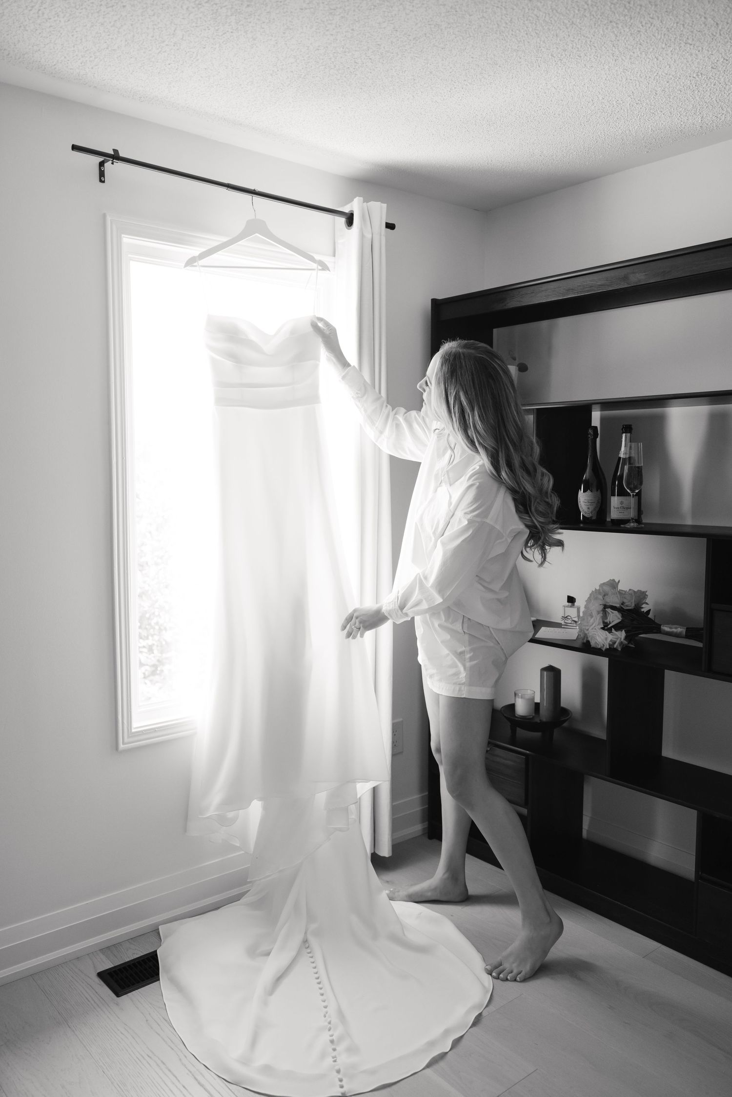 A white wedding dress hanging by a window during bridal preparation.